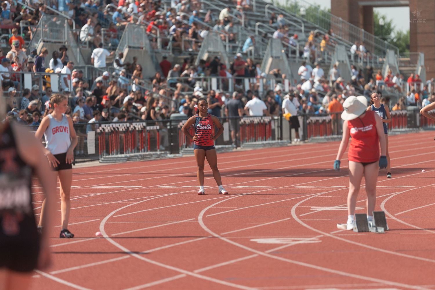 Athletes stand on a red track during an outdoor track and field competition with packed stadium bleachers.