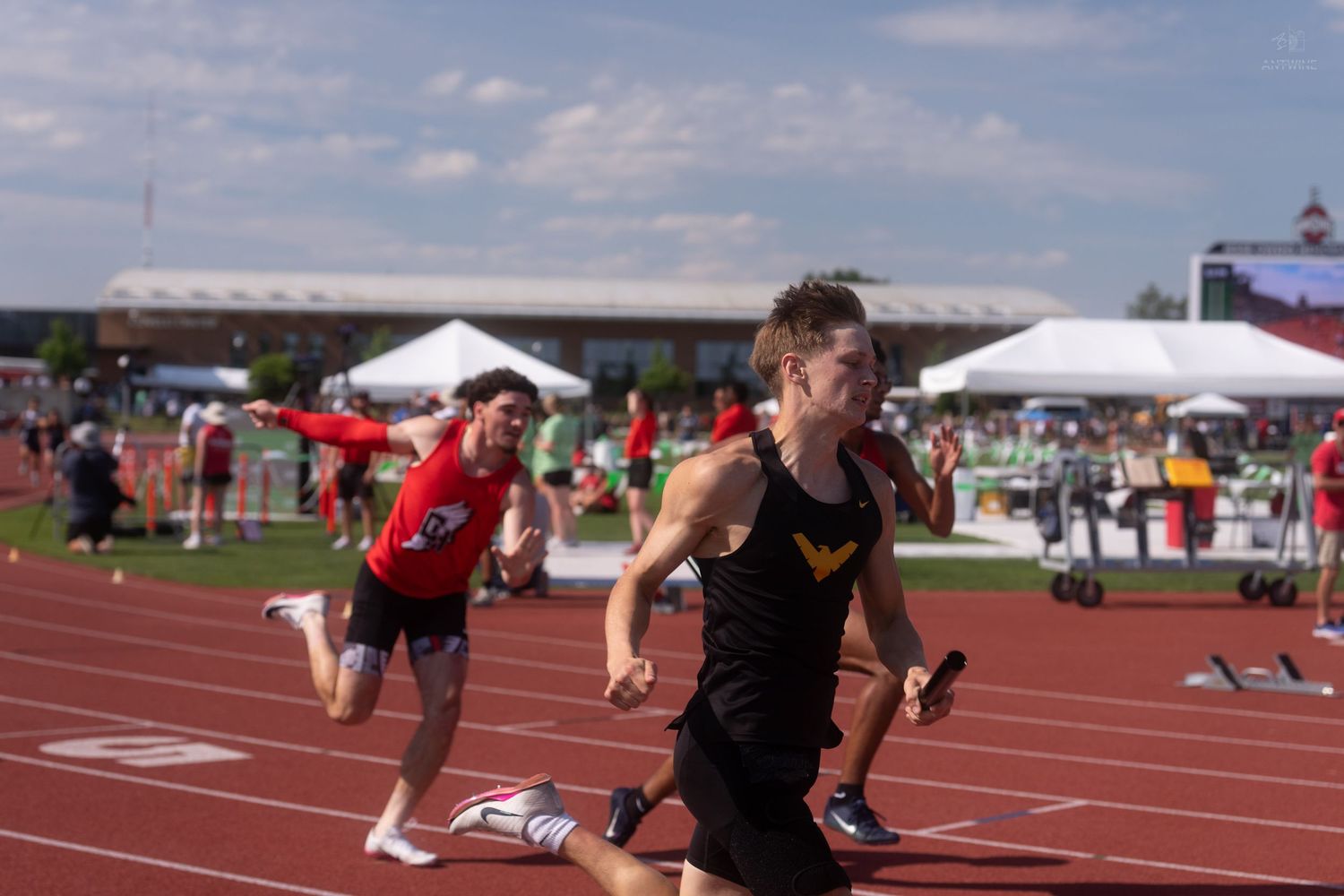 Young athlete in black uniform runs with a baton during a relay race at an outdoor track and field competition.