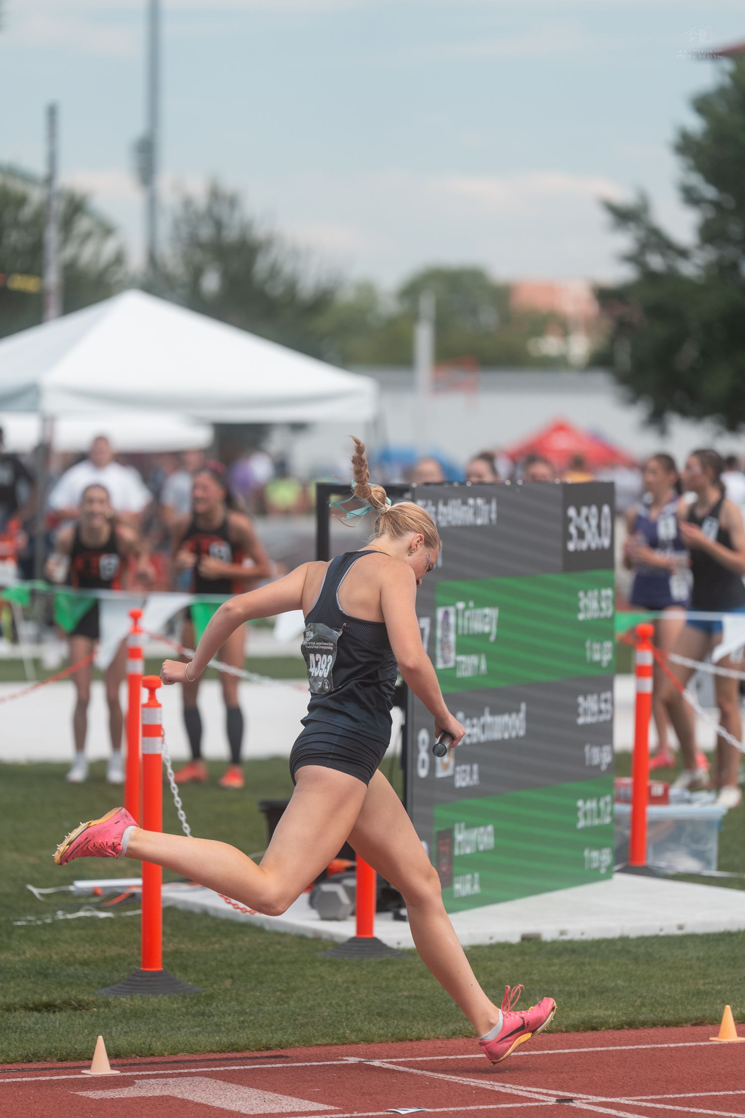 Female relay runner in black uniform crosses finish line at high school state track championship.
