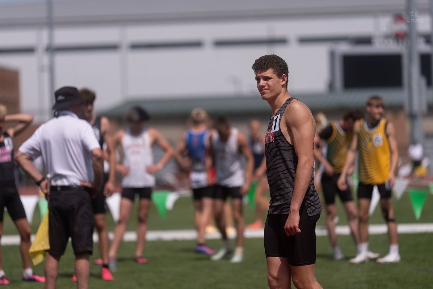 Male track athlete in dark uniform standing on infield during an outdoor track and field meet.