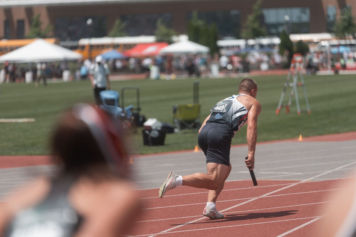 Track athlete running relay race carrying baton on red outdoor track during sunny athletic competition.