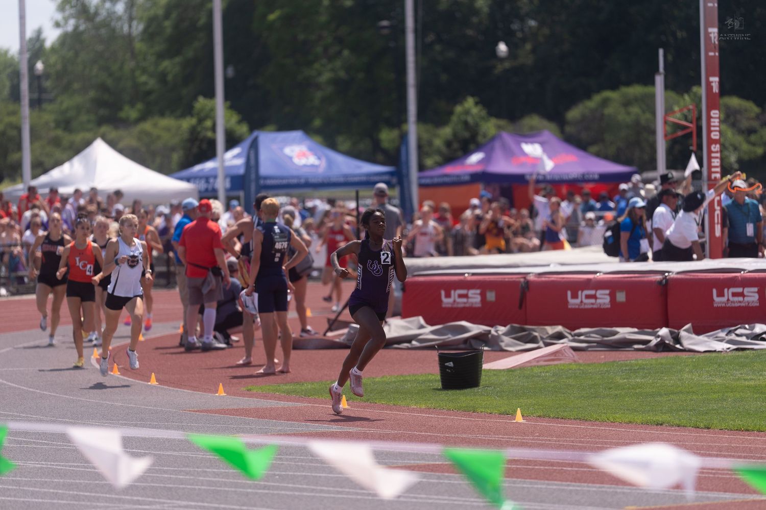 Male athlete sprinting on track at crowded outdoor collegiate track and field meet.