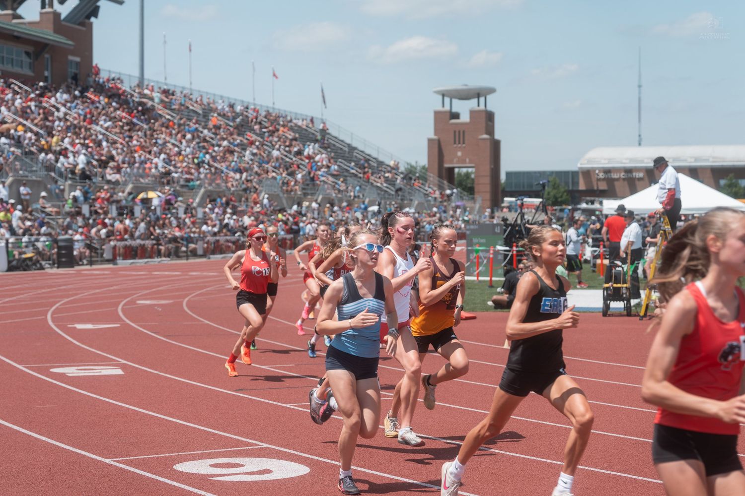Female athletes race around the track during a sunny high school track and field competition at a packed stadium.
