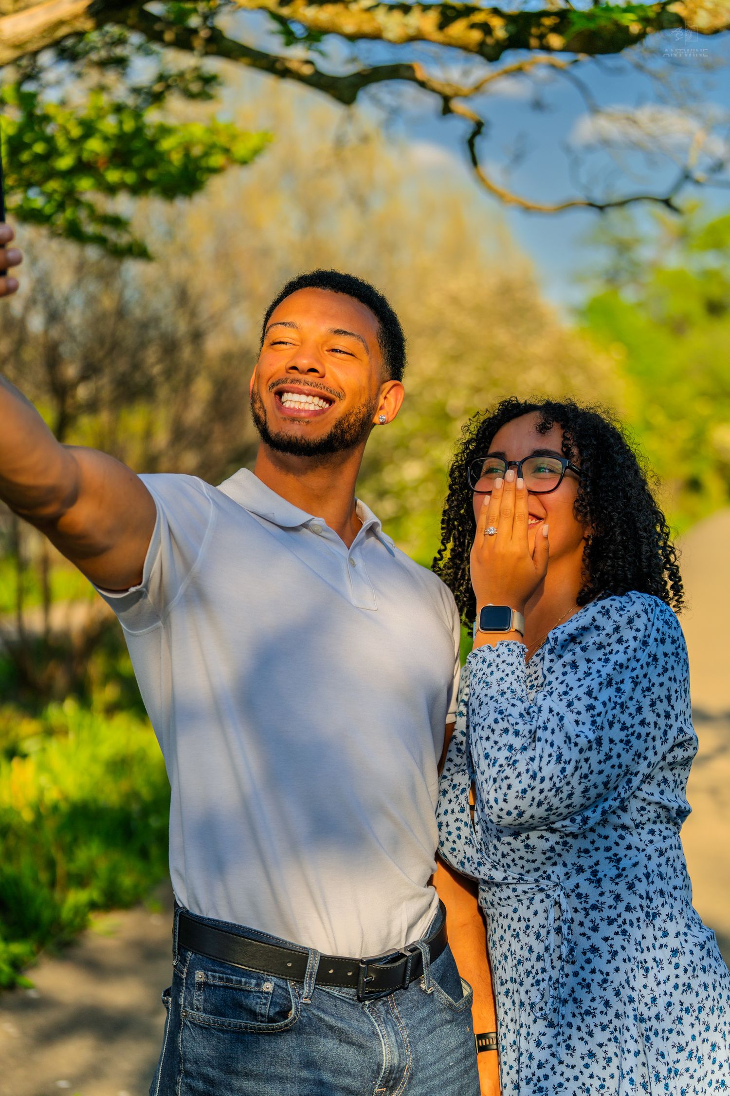 Couple taking a selfie in a sunny park, woman covering mouth with hand in joyful surprise.