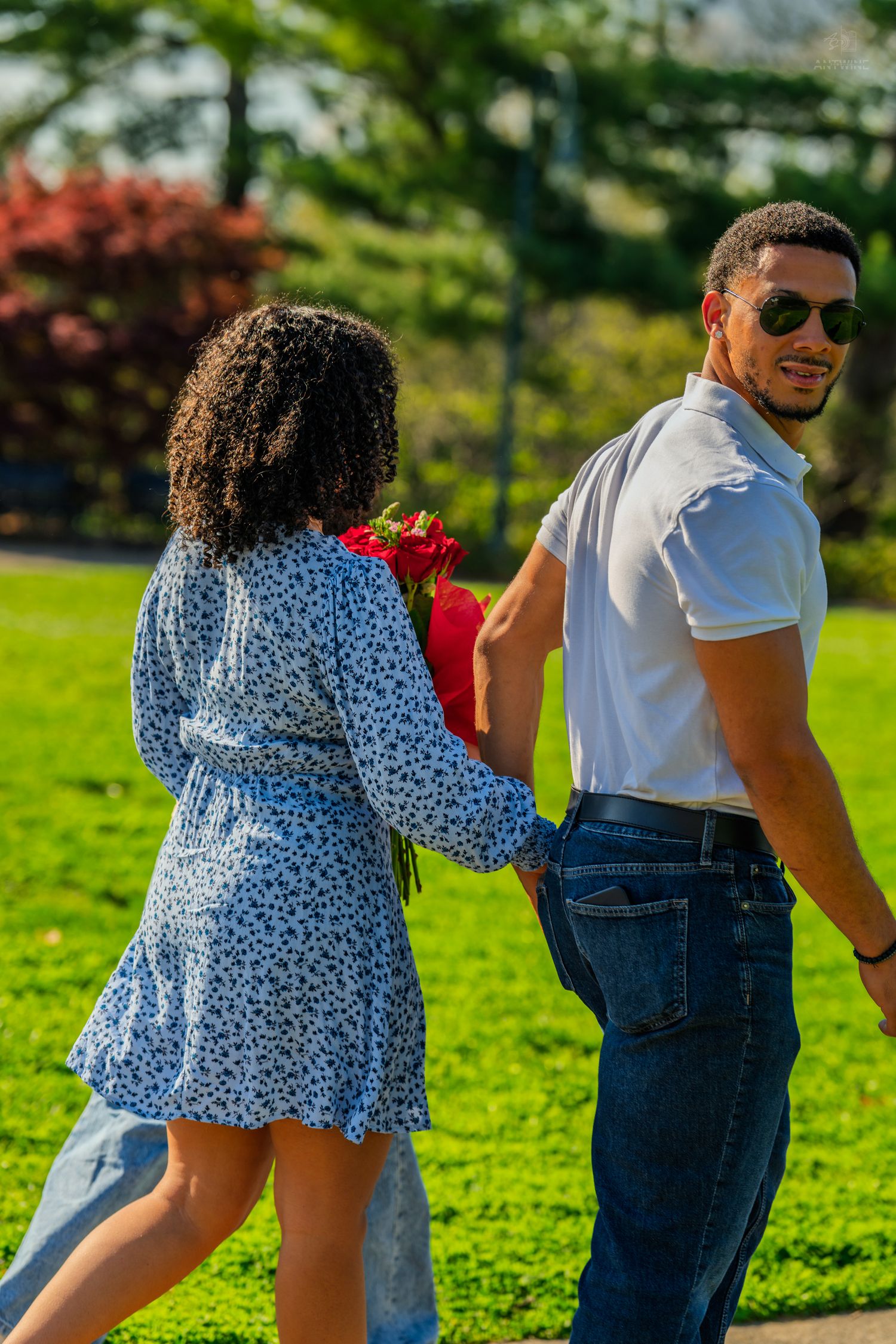 A couple walks hand in hand in a sunny park, woman in blue floral dress holding red roses.