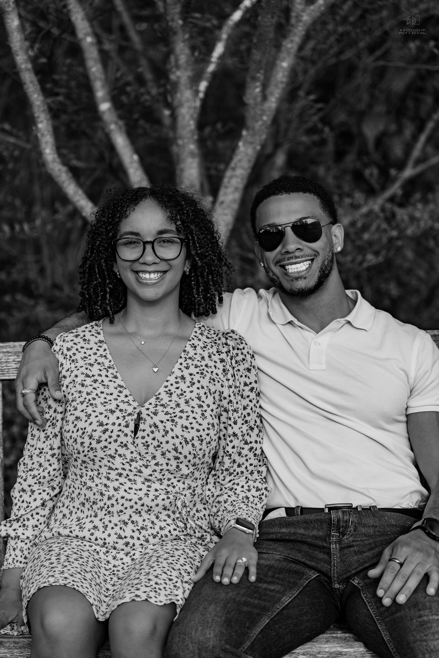 Black and white portrait of a smiling couple sitting on a bench with trees in the background.
