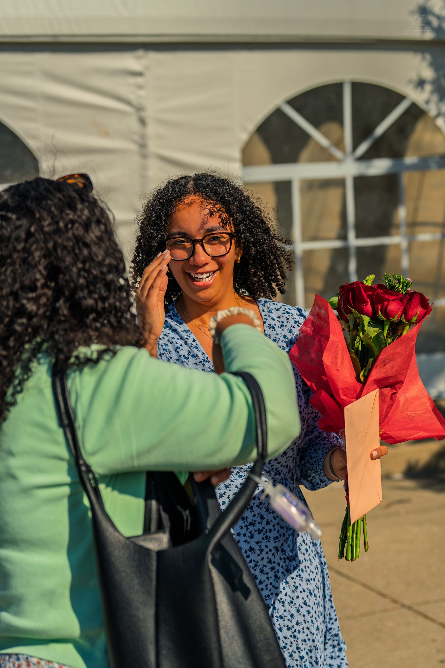 A joyful woman holding red roses and a card smiles as someone touches her cheek outdoors.