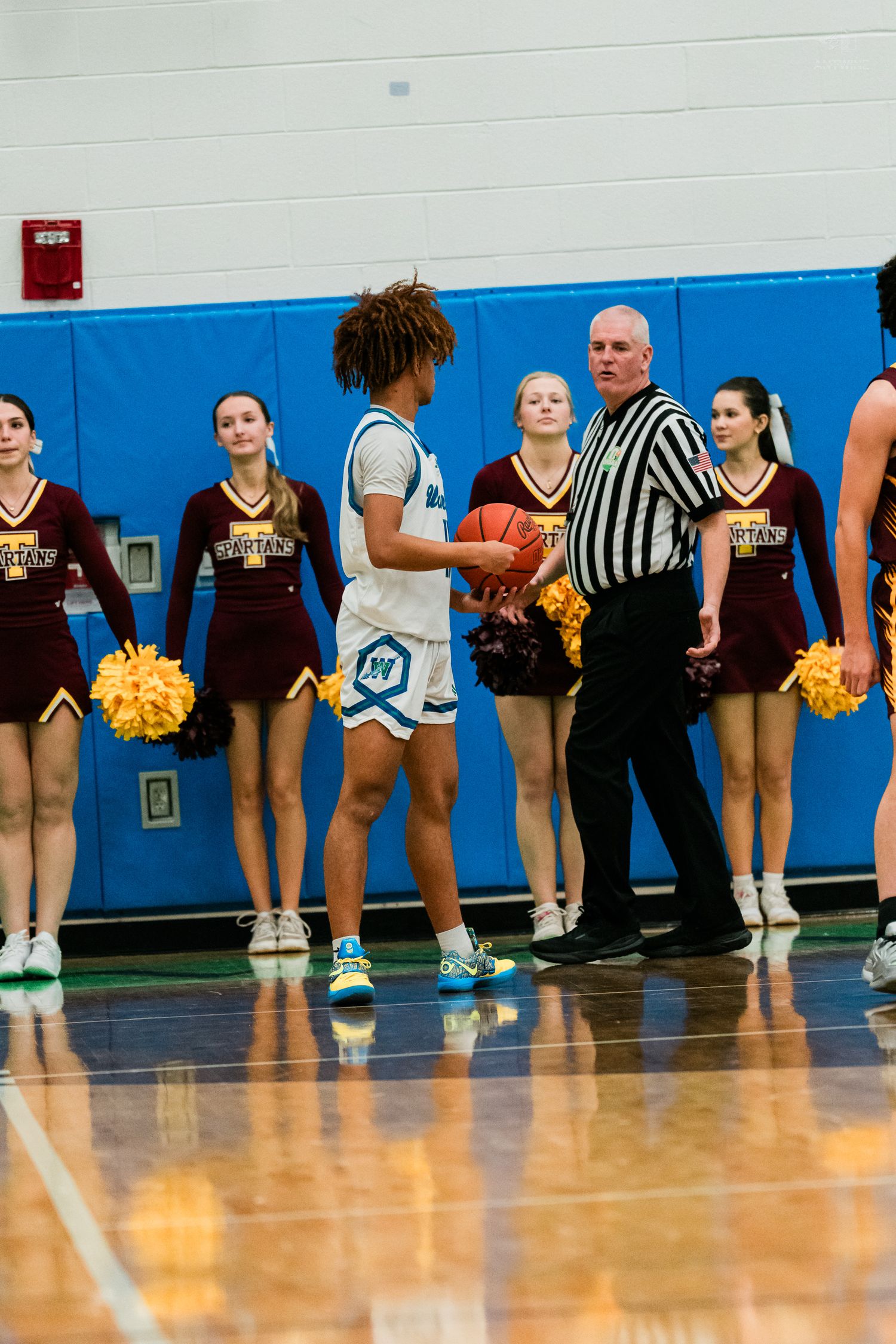 Basketball player receives ball from referee at sideline with cheerleaders in maroon uniforms in background.