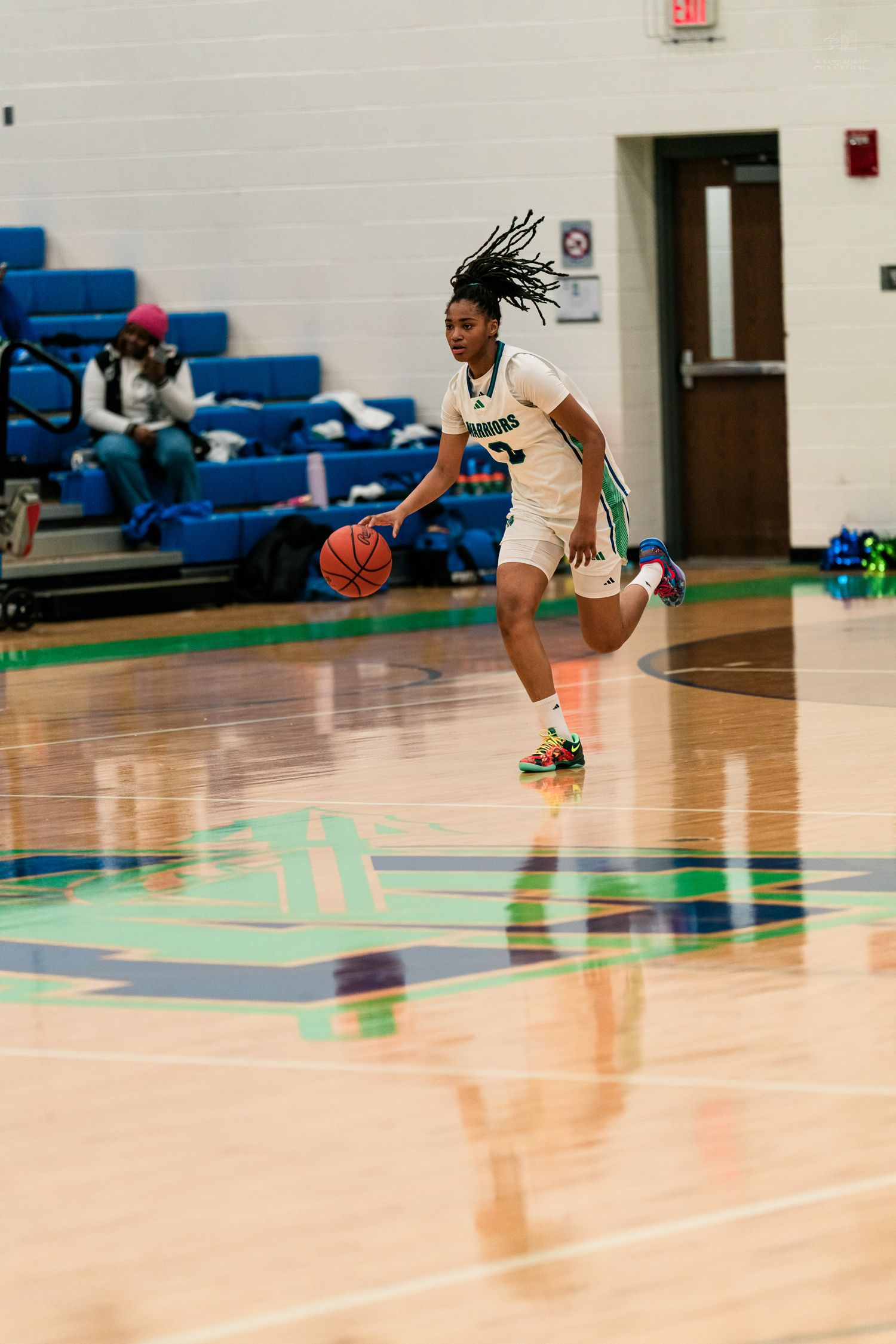 Female Warriors basketball player dribbles ball upcourt in teal uniform during high school game.