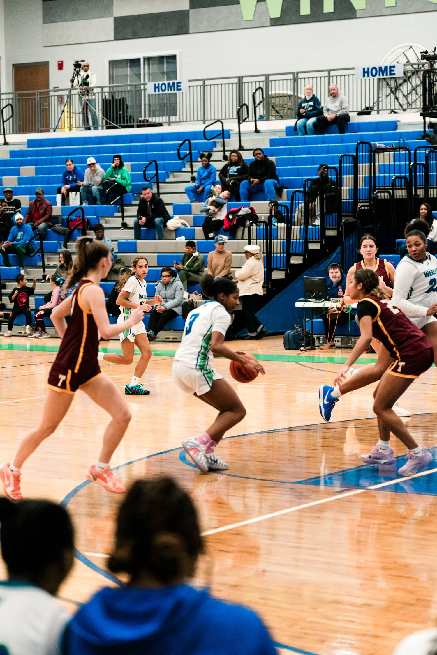 High school girls basketball player in teal dribbles through maroon defenders during a game in a blue and white gymnasium.
