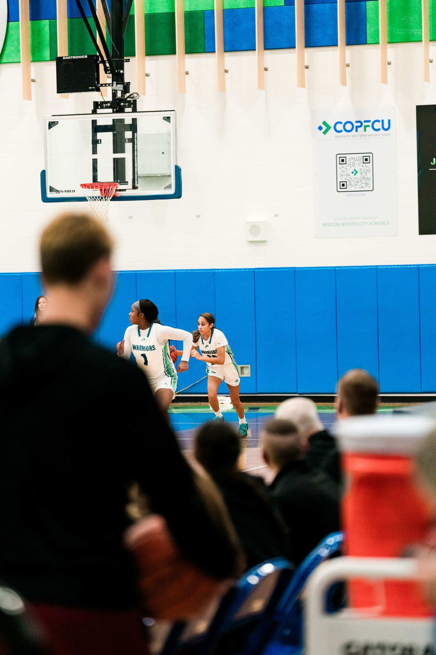 Two Warriors basketball players advance the ball upcourt as spectators watch from the sideline in a high school gymnasium.
