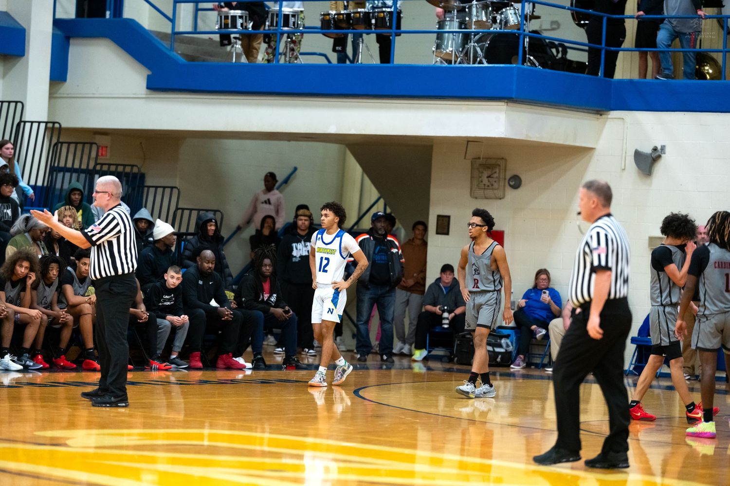 Two referees and a player in white jersey number 12 stand on a high school basketball court during a game.