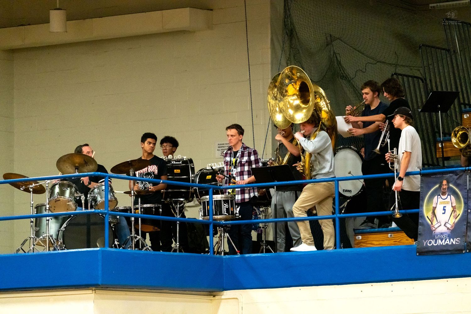 High school pep band with tubas and drums performs on a blue balcony during a basketball game.