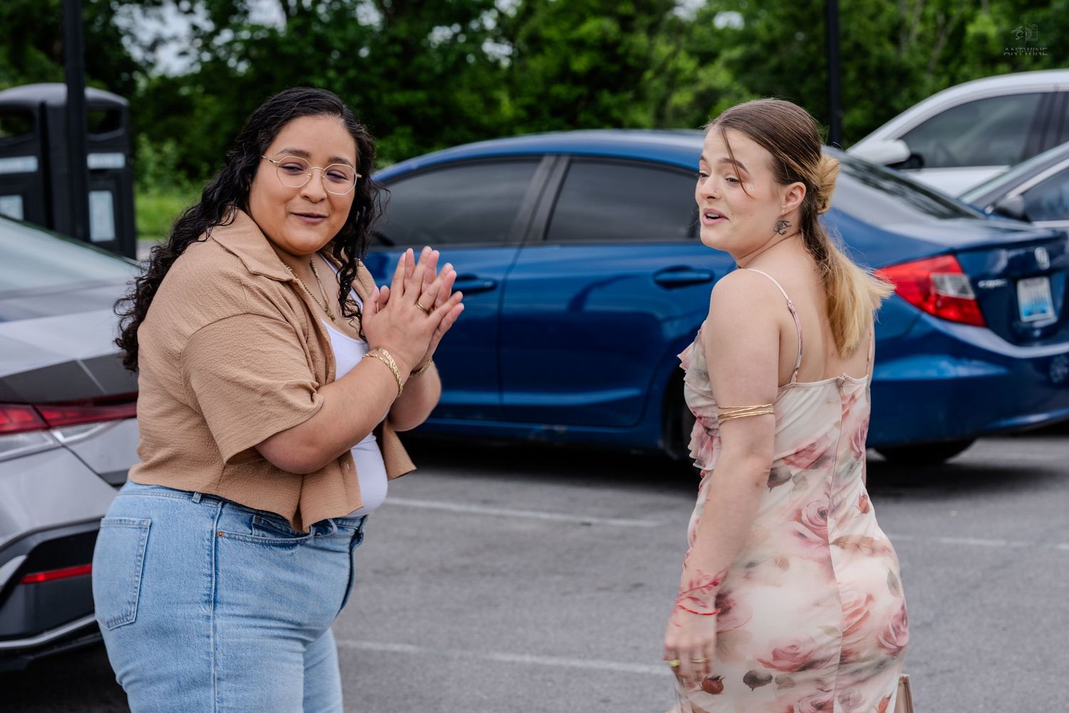 Two women face each other laughing and clapping in a parking lot near colorful cars.