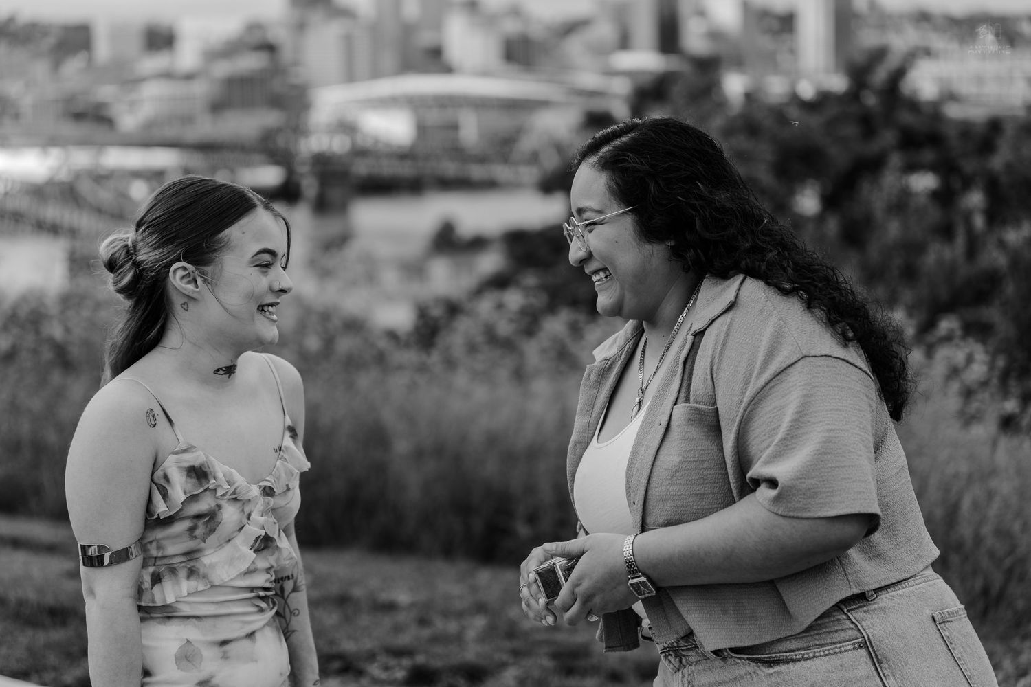 Black and white photo of two women smiling and conversing outdoors with cityscape behind.