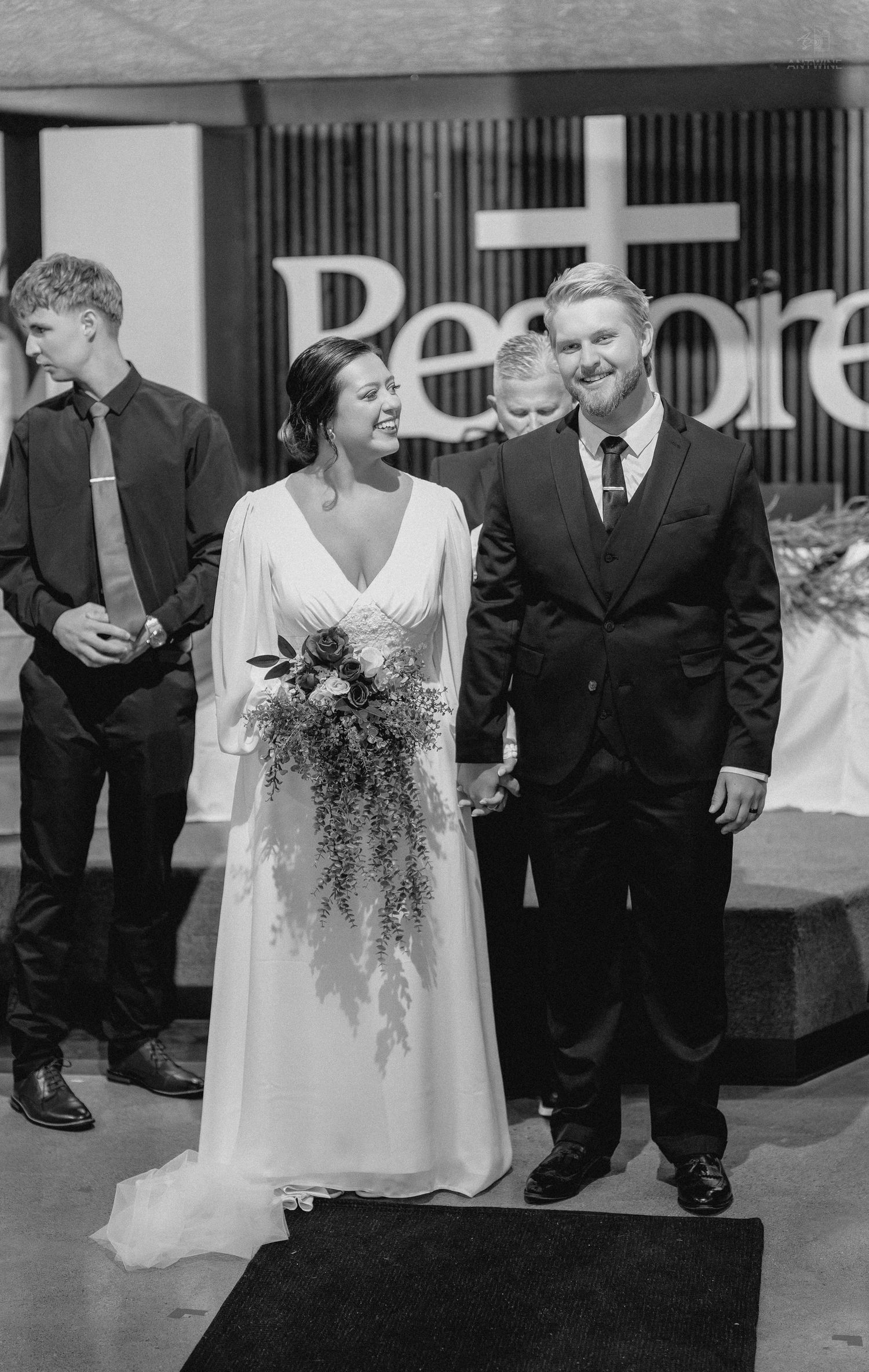 Black and white wedding photo of smiling bride in white gown holding bouquet with groom in suit at church altar.