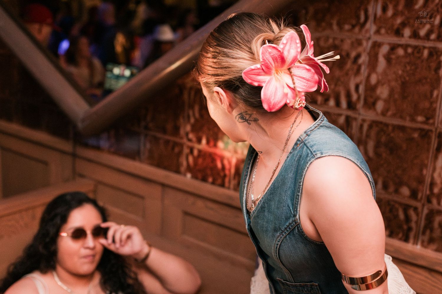 Woman with pink flower hair accessory and denim vest at a rustic indoor venue.