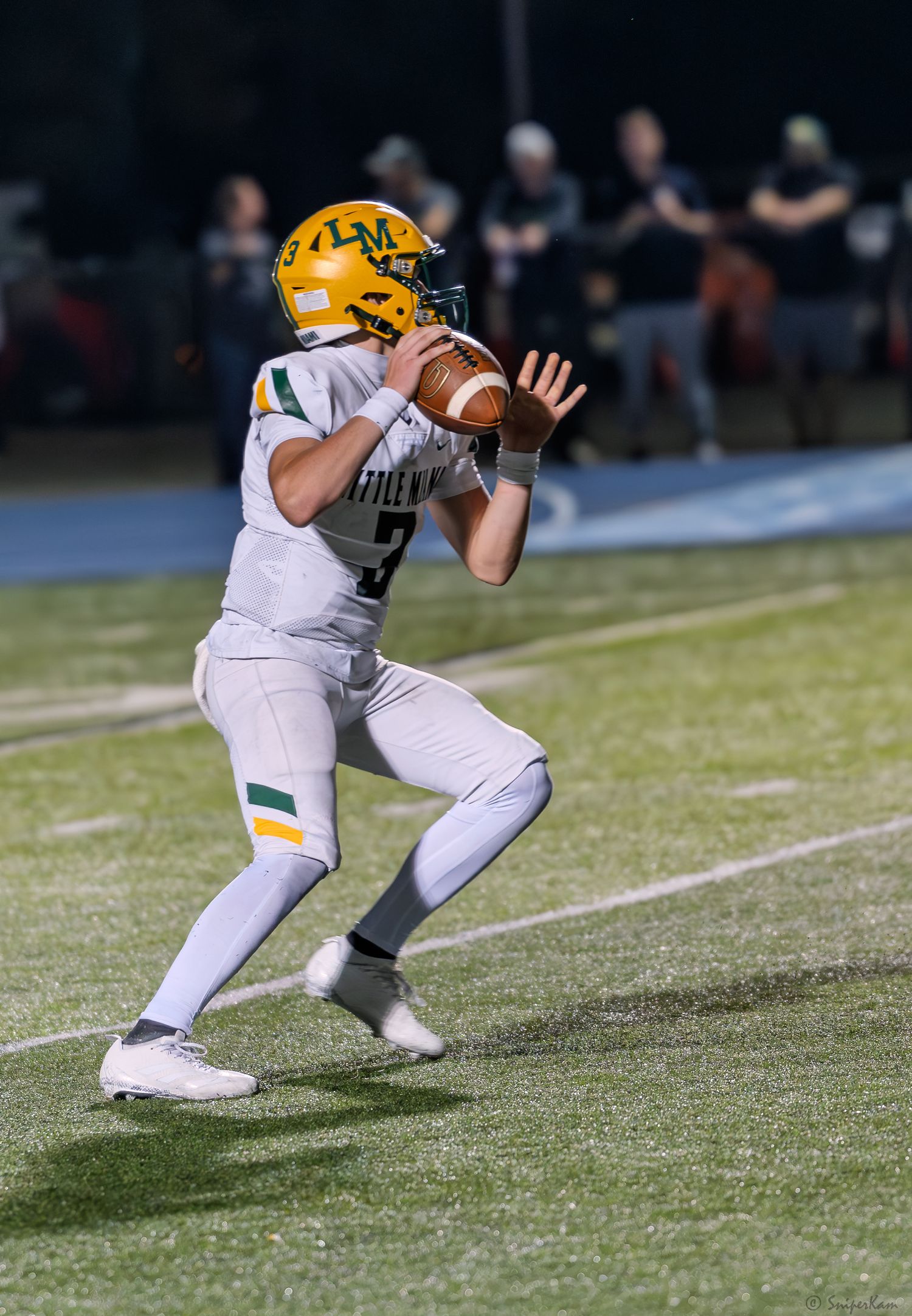 A quarterback in yellow and white uniform drops back to pass during a night football game.