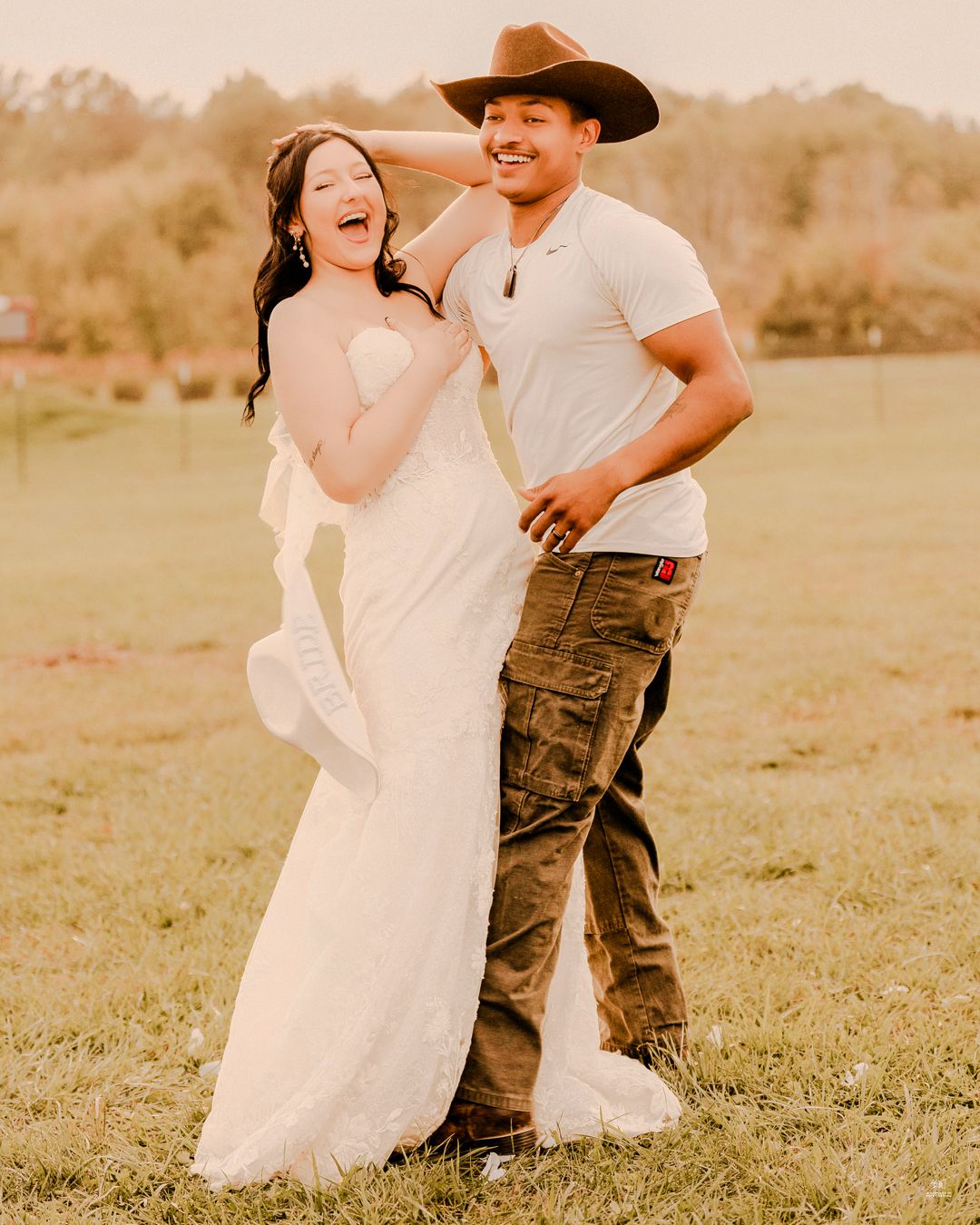 Laughing bride in a white gown and groom in a cowboy hat share a playful moment on a lawn.