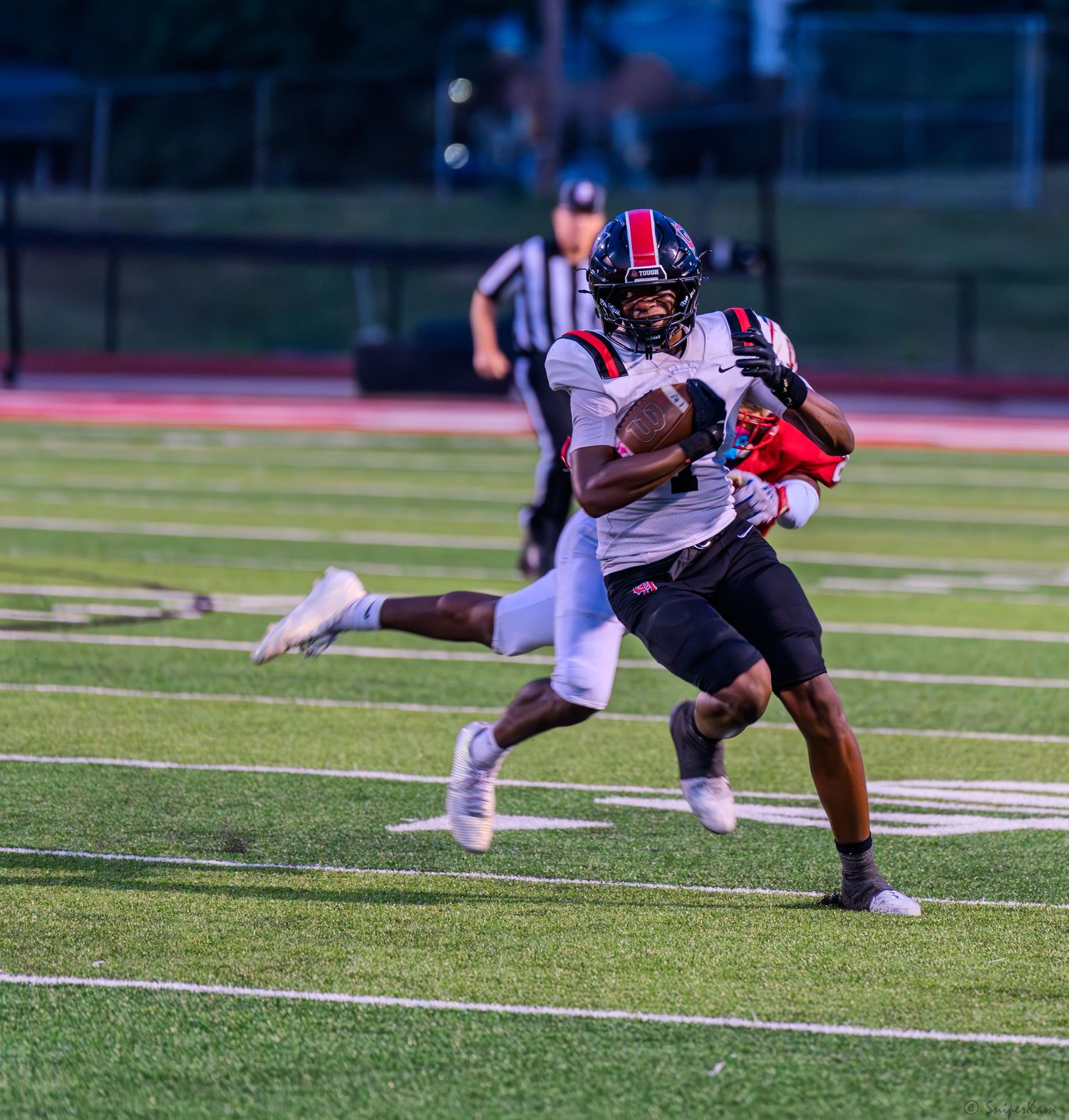 High school football player in dark uniform runs with ball during evening game on turf.