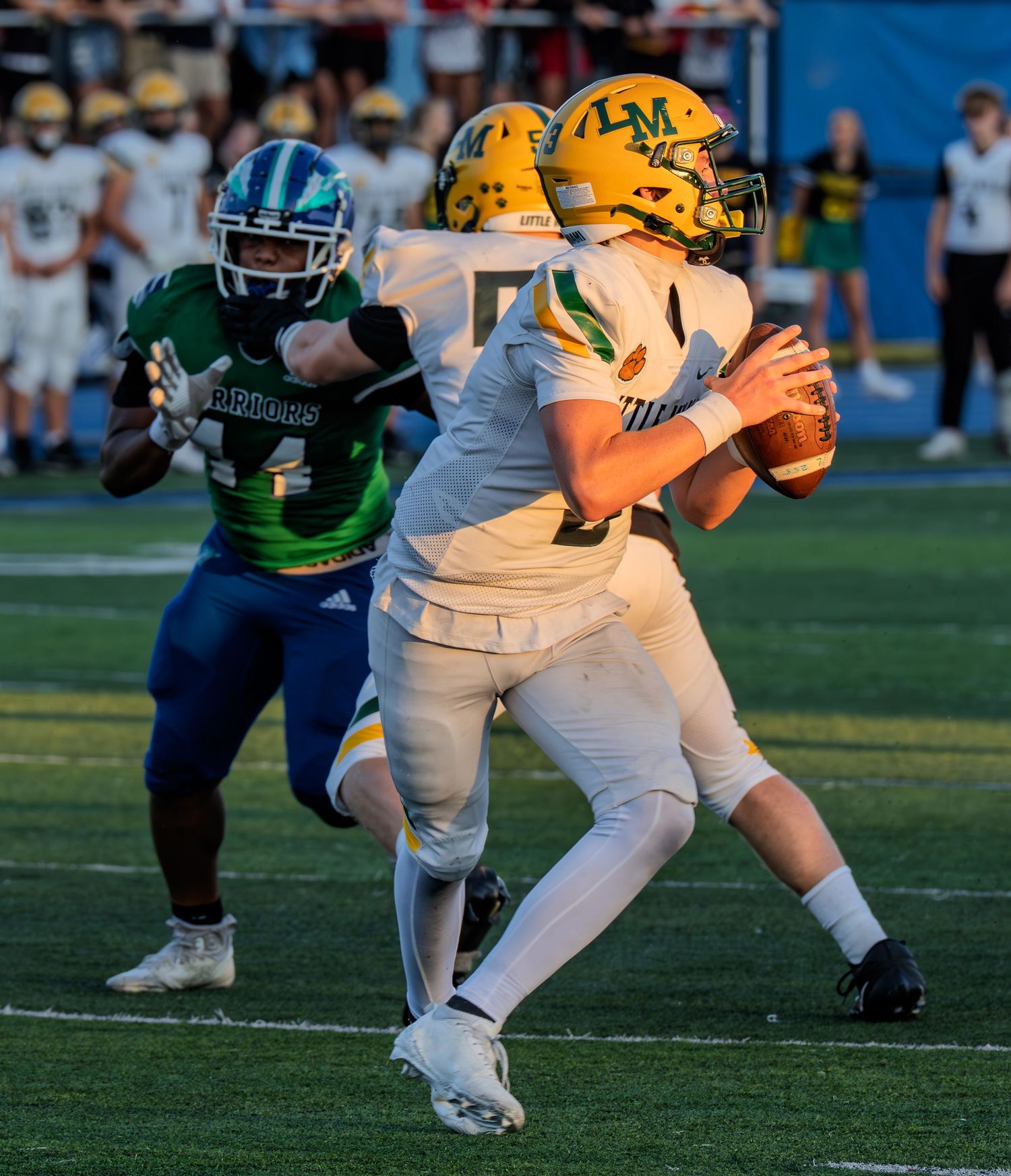 High school quarterback in white and gold uniform scrambles while being pursued by a defender in green.