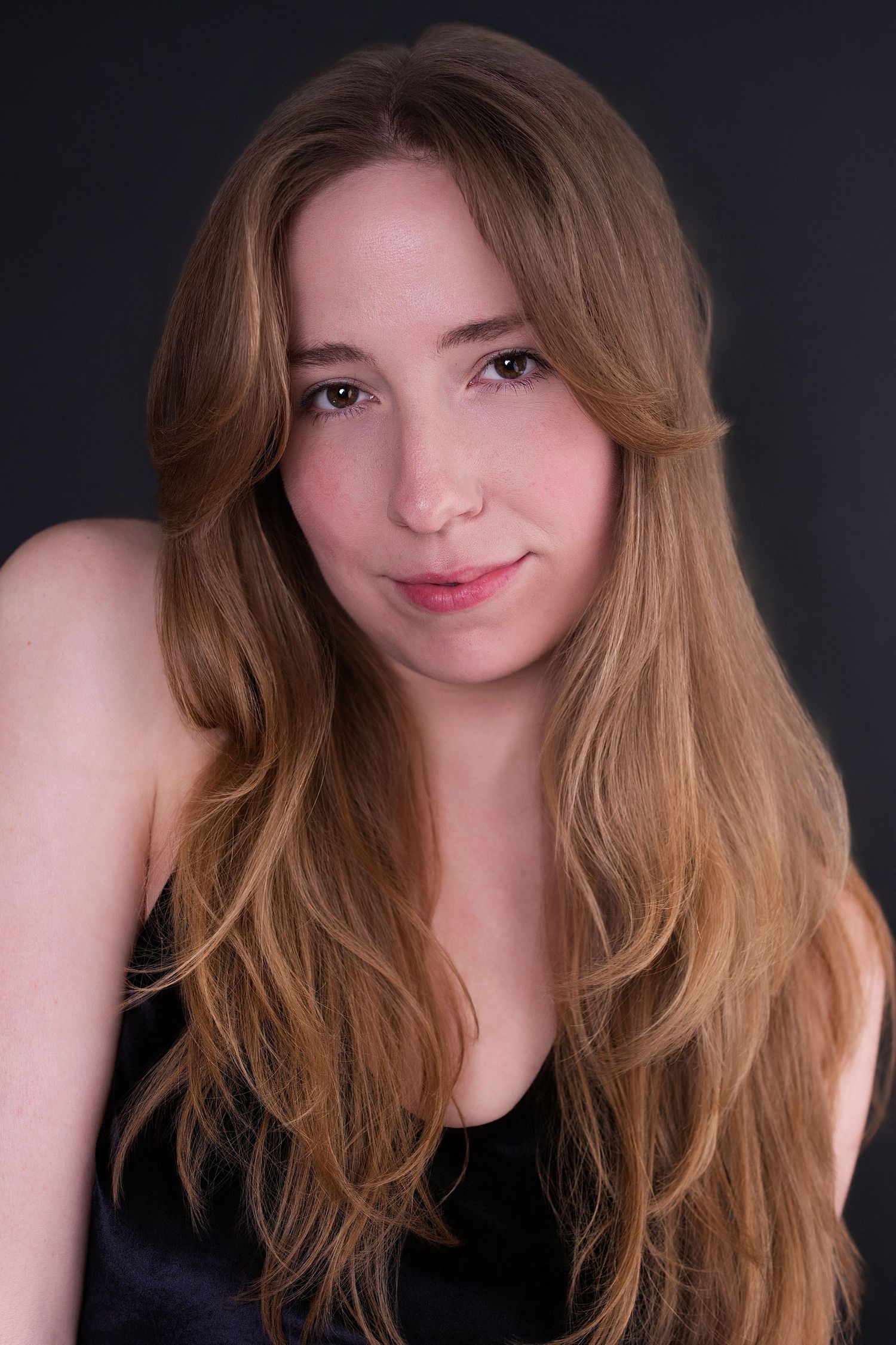 Young woman with long auburn wavy hair wearing black top, smiling gently against dark gray background.