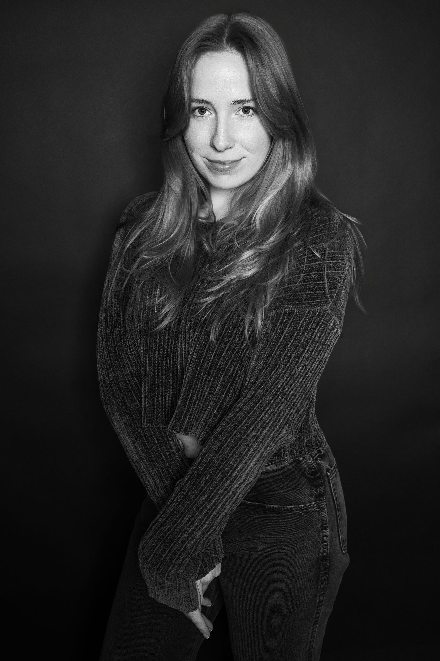 Black and white studio portrait of a young woman in a dark knit sweater and jeans posing confidently.