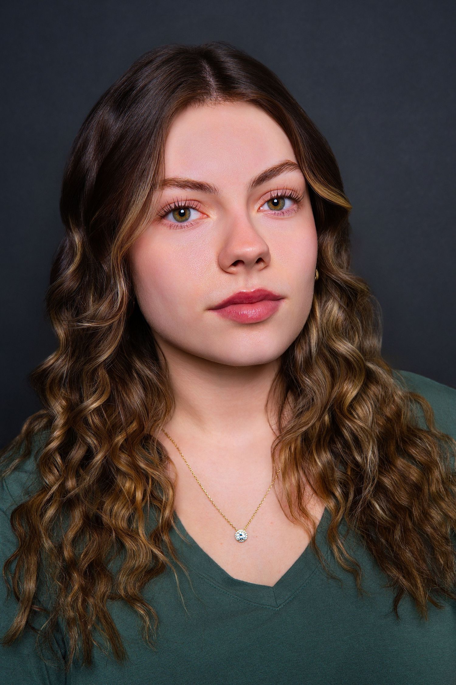 Young woman with long curly highlighted hair wearing green v-neck top and gold necklace.