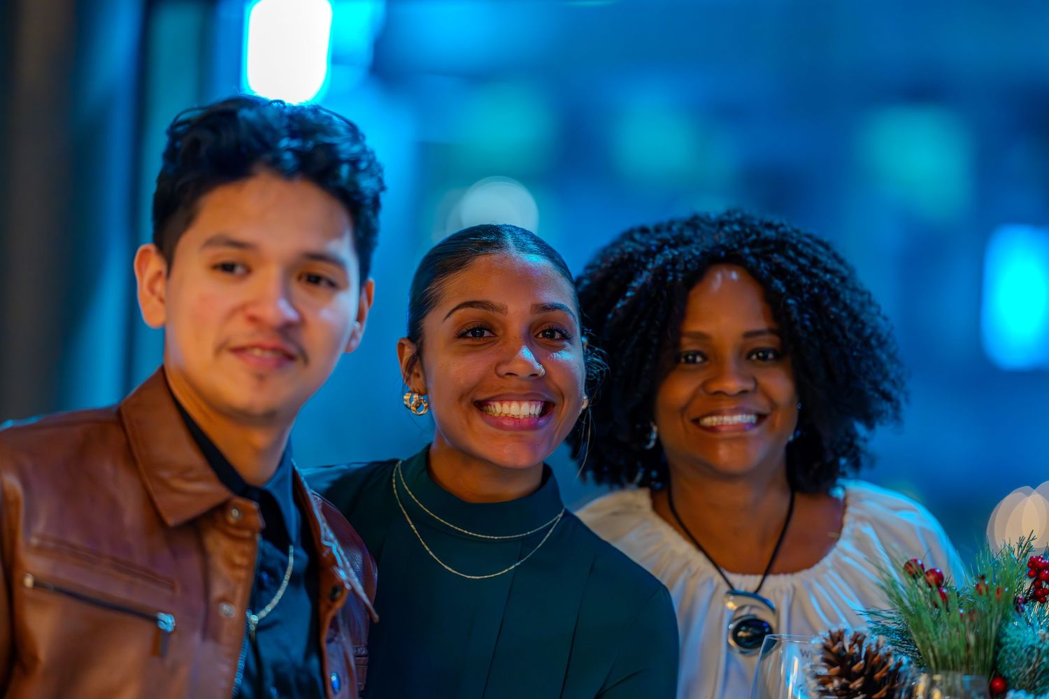 Three smiling friends pose together at a festive holiday event with blue bokeh lights in the background.