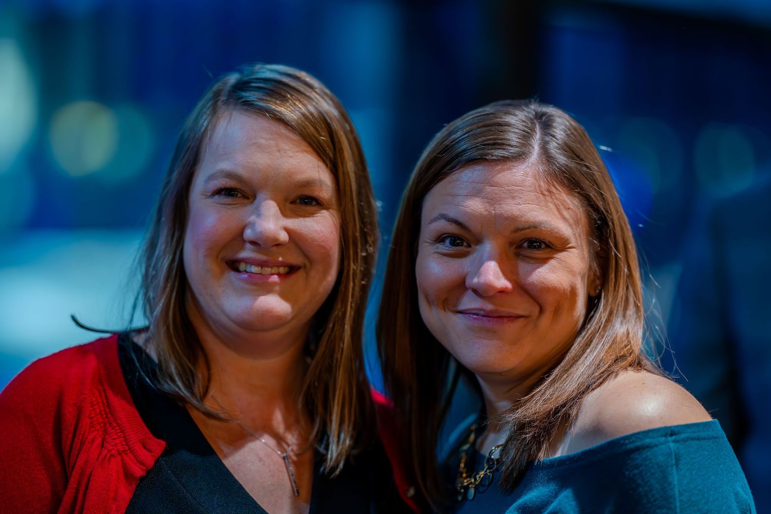 Two smiling women pose together at an indoor event, one in red and one in teal off-shoulder top.