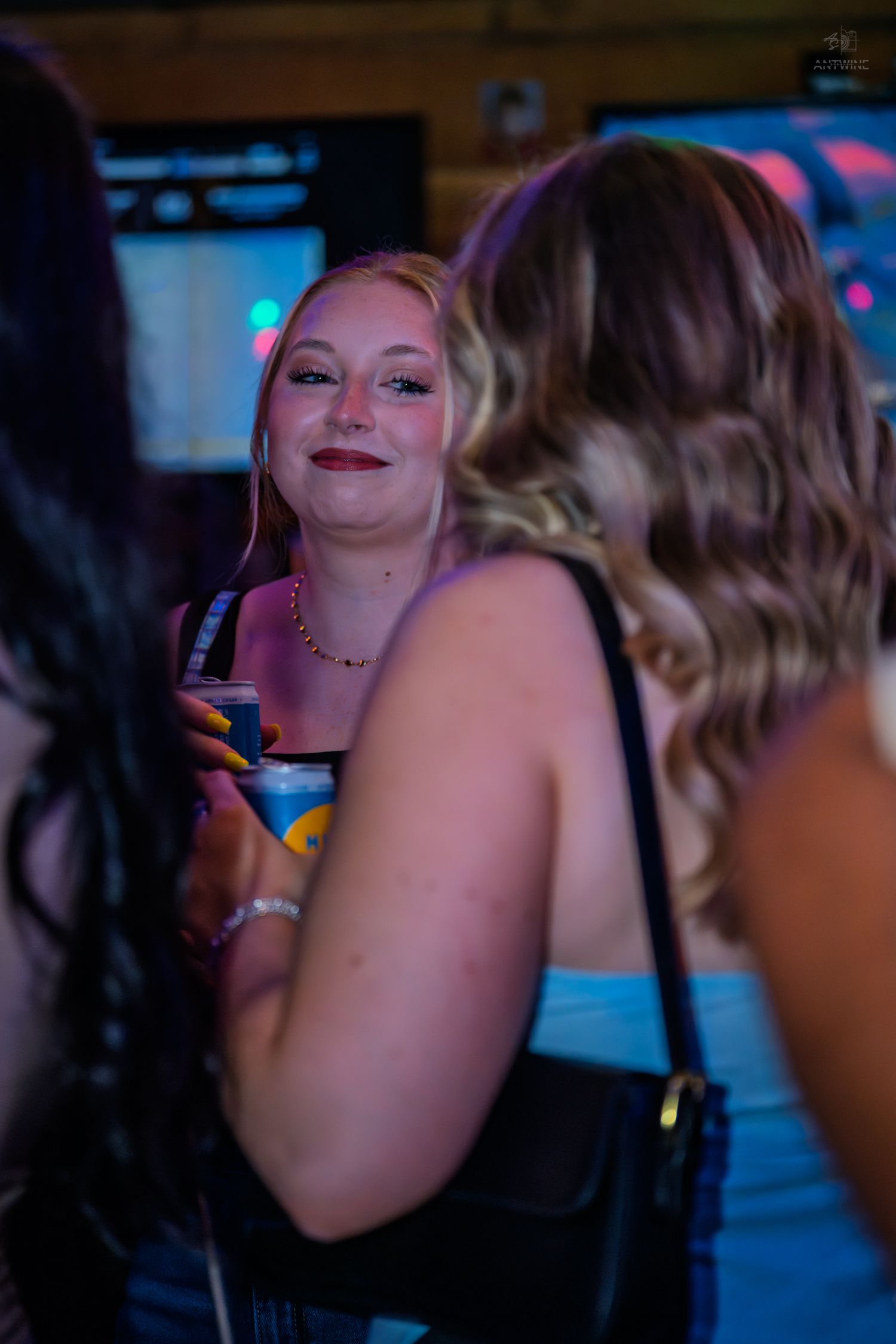 Young woman smiling and holding a cocktail at a nightclub, chatting with friends in a dimly lit bar.