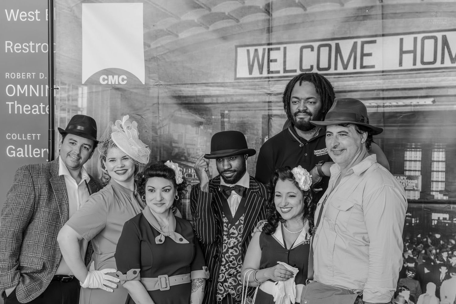 Group of seven people in 1940s vintage costumes posing under a Welcome Home sign in black and white photo.