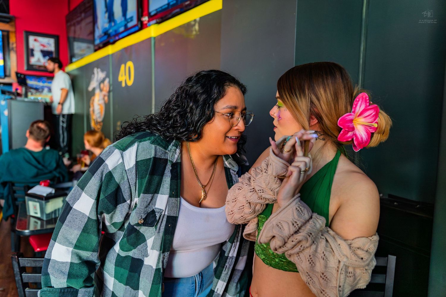 Two women smiling at each other at a bar, one wearing a floral hair accessory and green top.