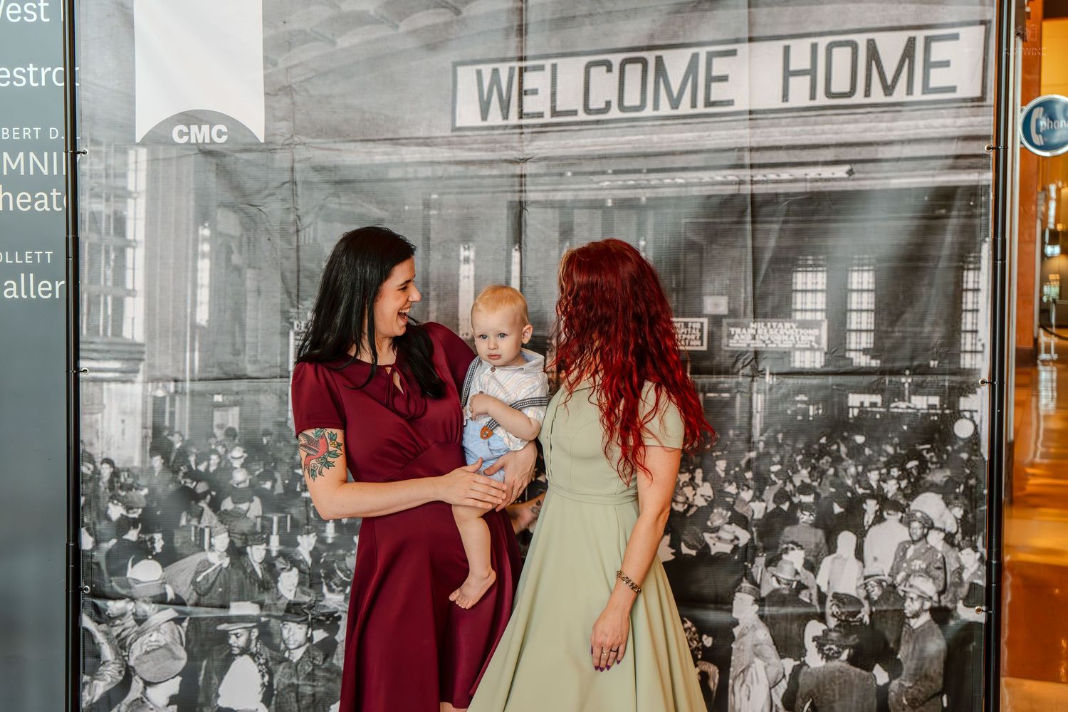 Two women and a baby pose in front of a Welcome Home backdrop at a museum exhibit.