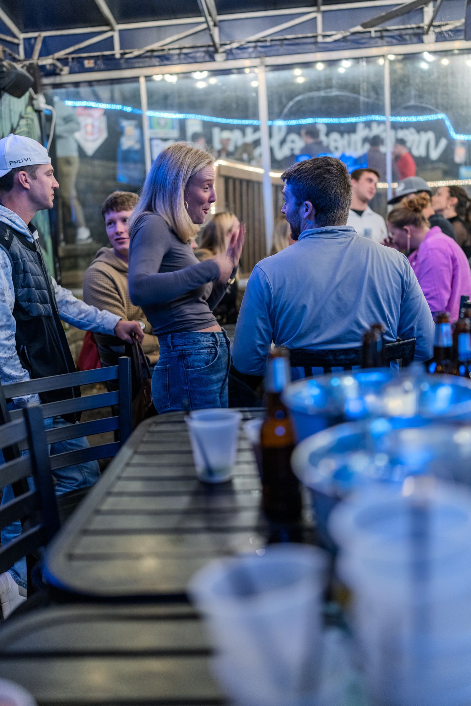 Young adults socializing at an outdoor bar patio with string lights, beer bottles, and plastic cups on the counter.