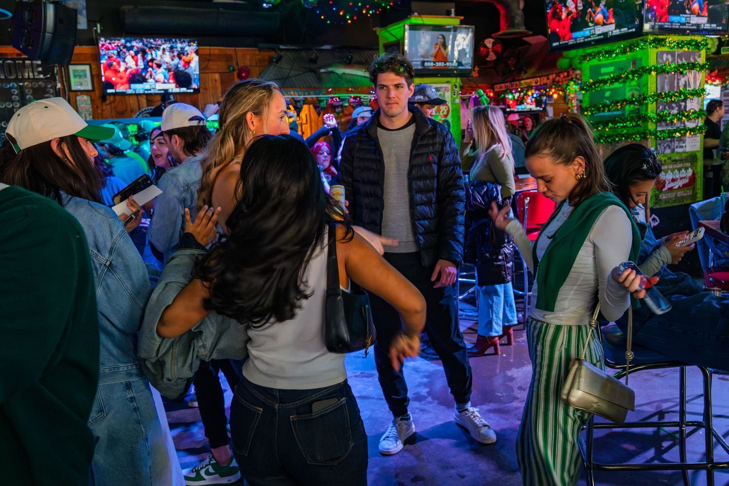 People dancing and socializing in a lively bar with neon green lights and TVs in the background.
