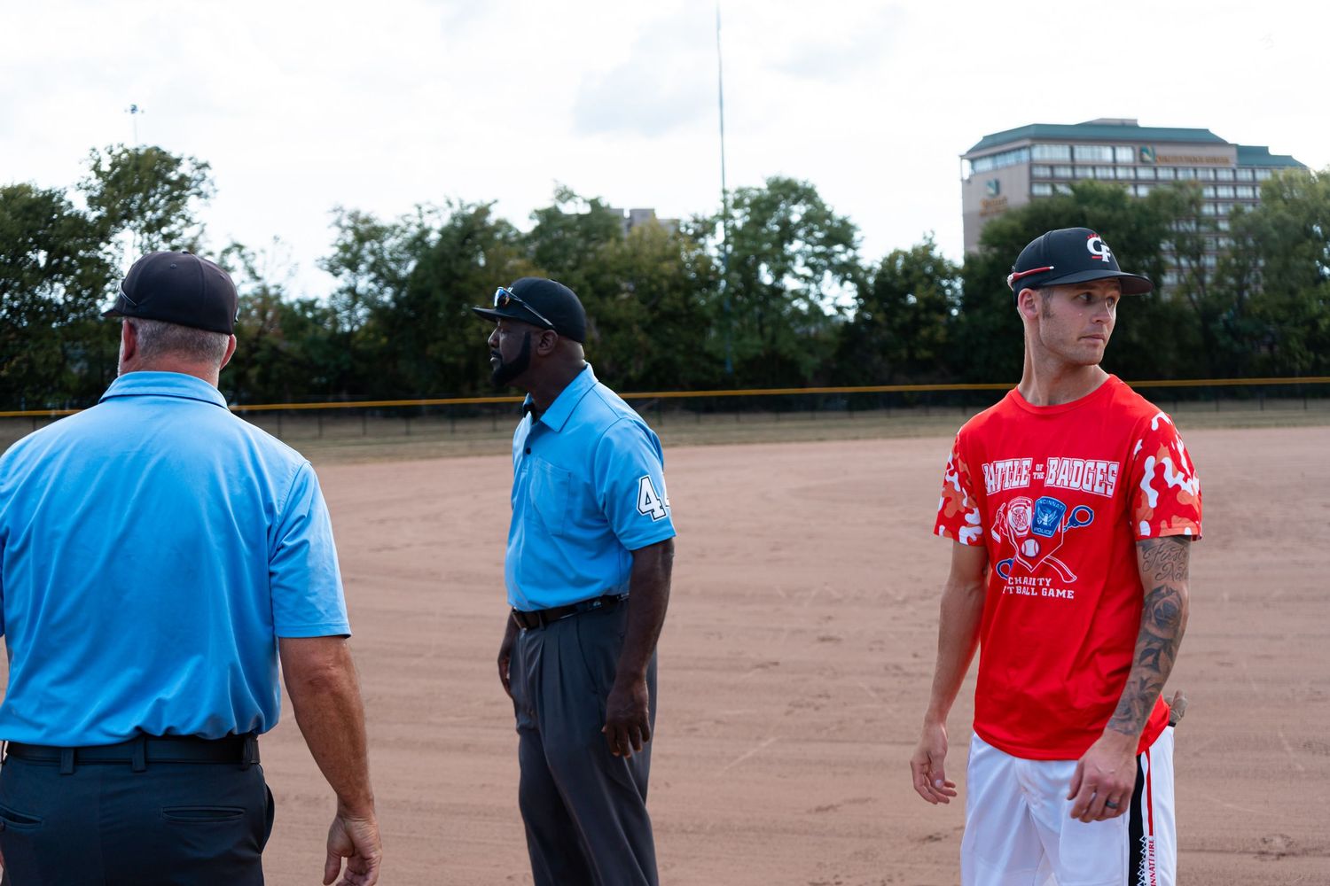 Two umpires in blue shirts stand with a player in a red jersey on a softball field.