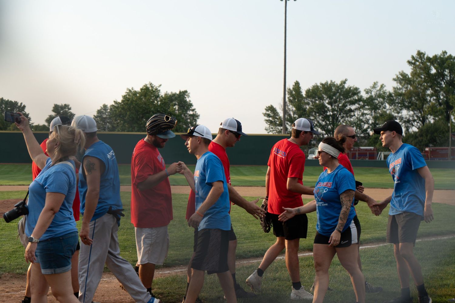 Teams shake hands after a charity softball game on a baseball field at dusk.