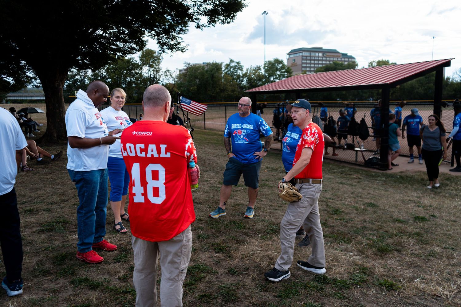 Group of people in sports jerseys, including Local 18, gather on a baseball field near a dugout for an outdoor event.
