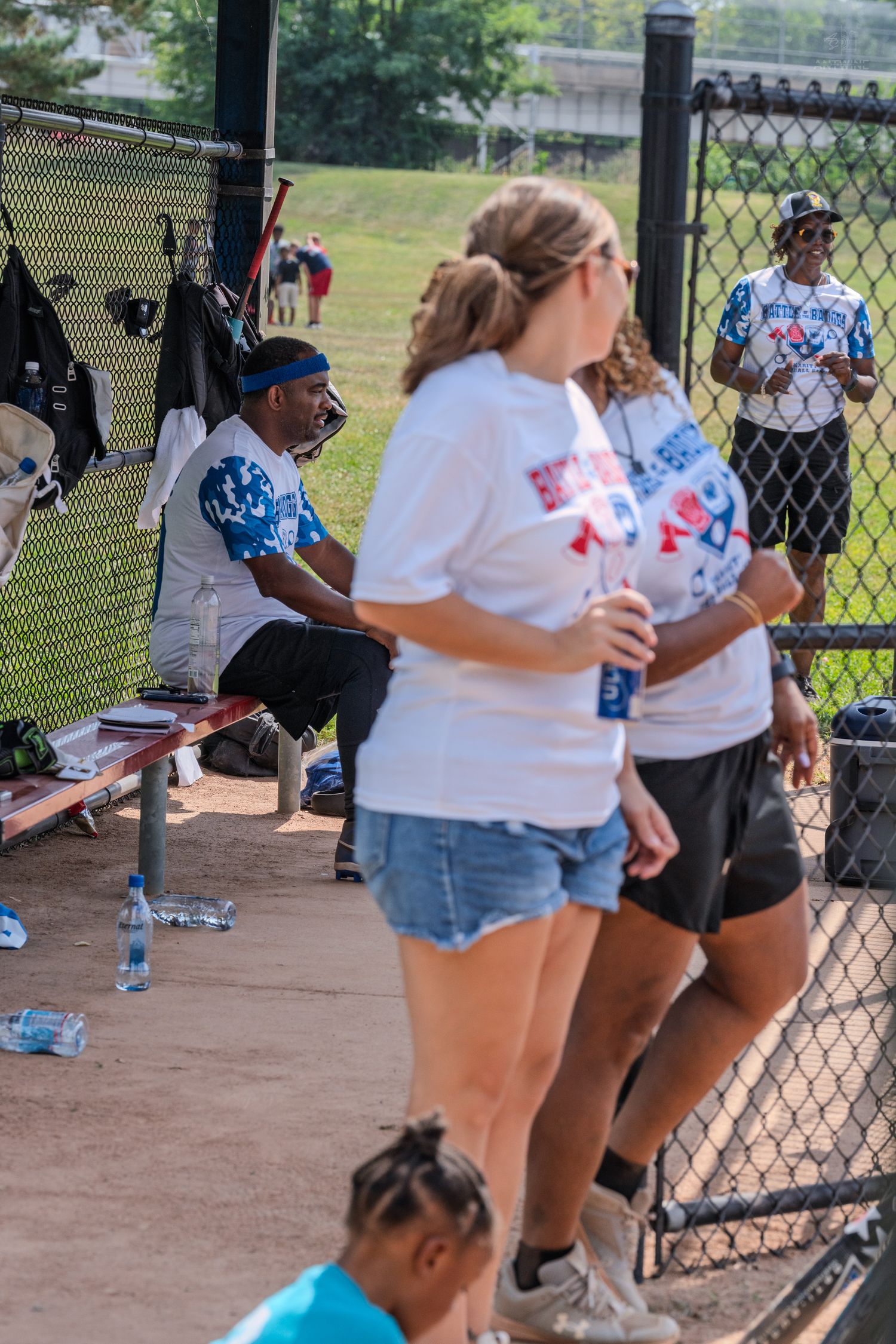 People gathered in a baseball dugout wearing white t-shirts during a sunny outdoor softball game event.