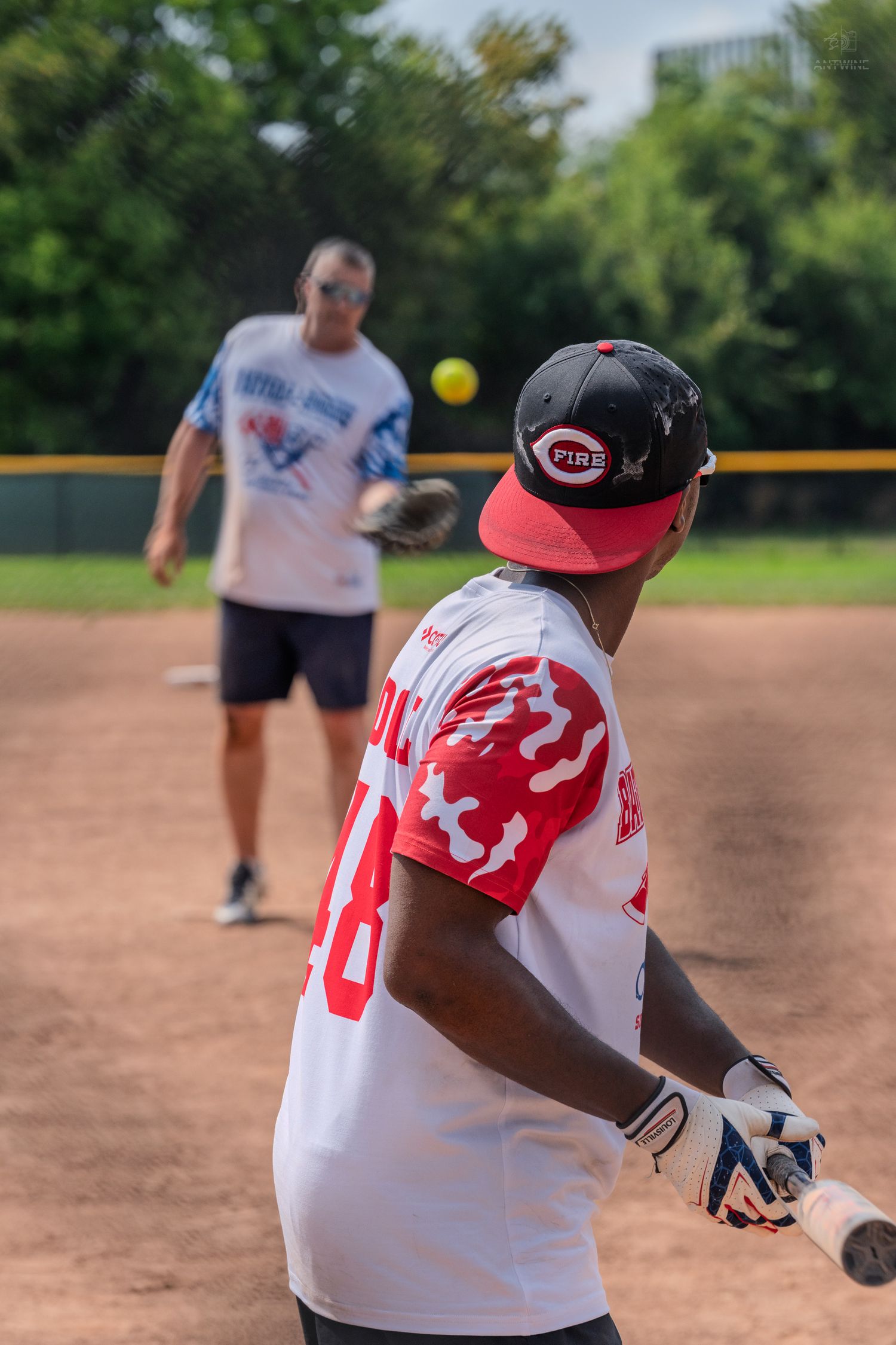 Batter in Reds jersey prepares to hit as pitcher throws yellow softball on outdoor dirt baseball field.