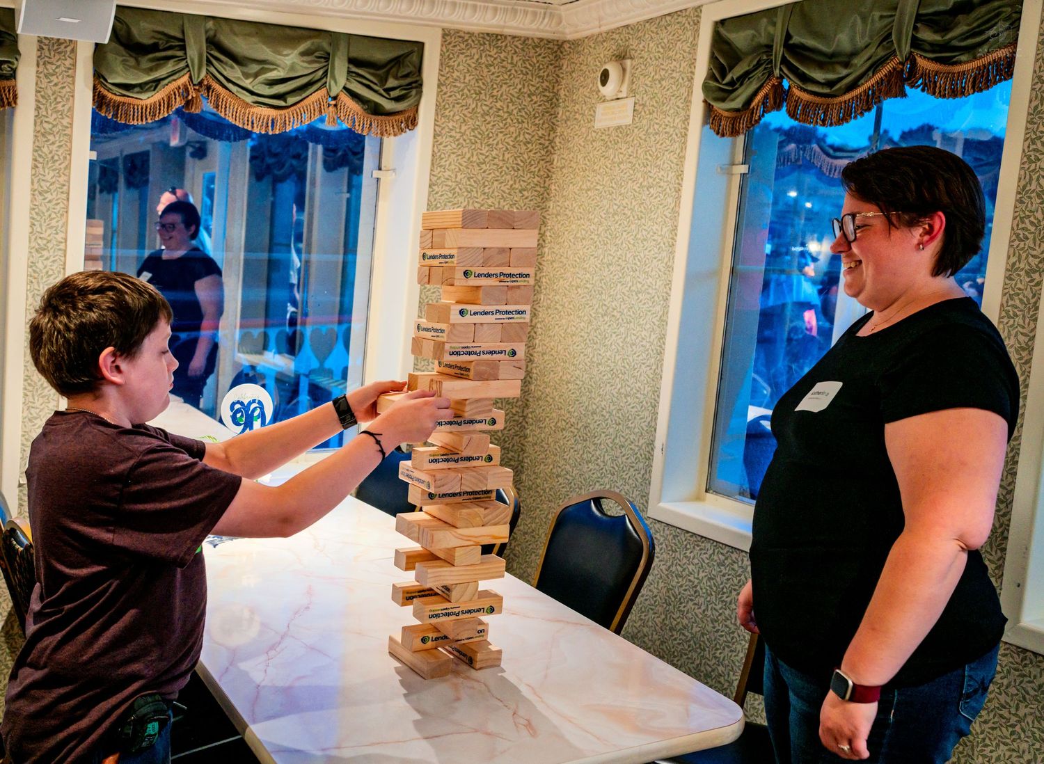Two people playing a giant Jenga tower game on a marble table indoors near large windows at night.