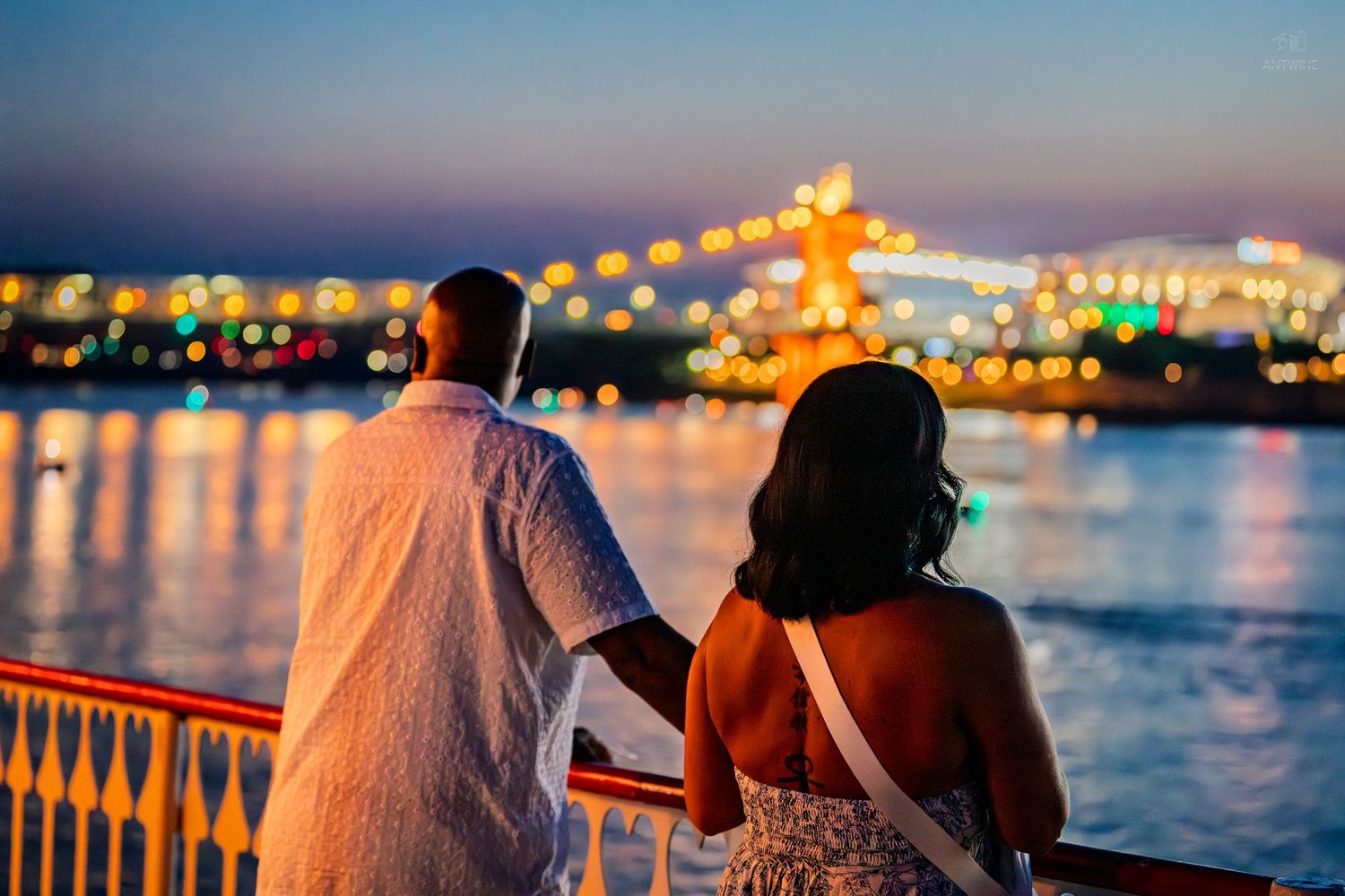 Couple holding hands on a waterfront railing enjoying a colorful city skyline at twilight.