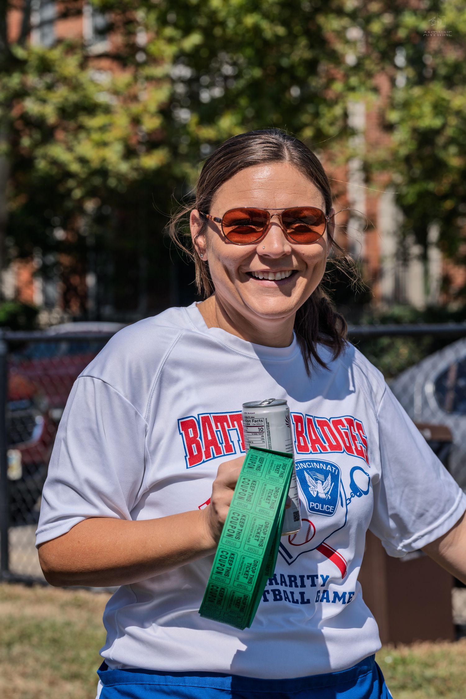 Smiling woman in sunglasses wearing a Battle Badges Cincinnati Police Charity Softball Game t-shirt outdoors.