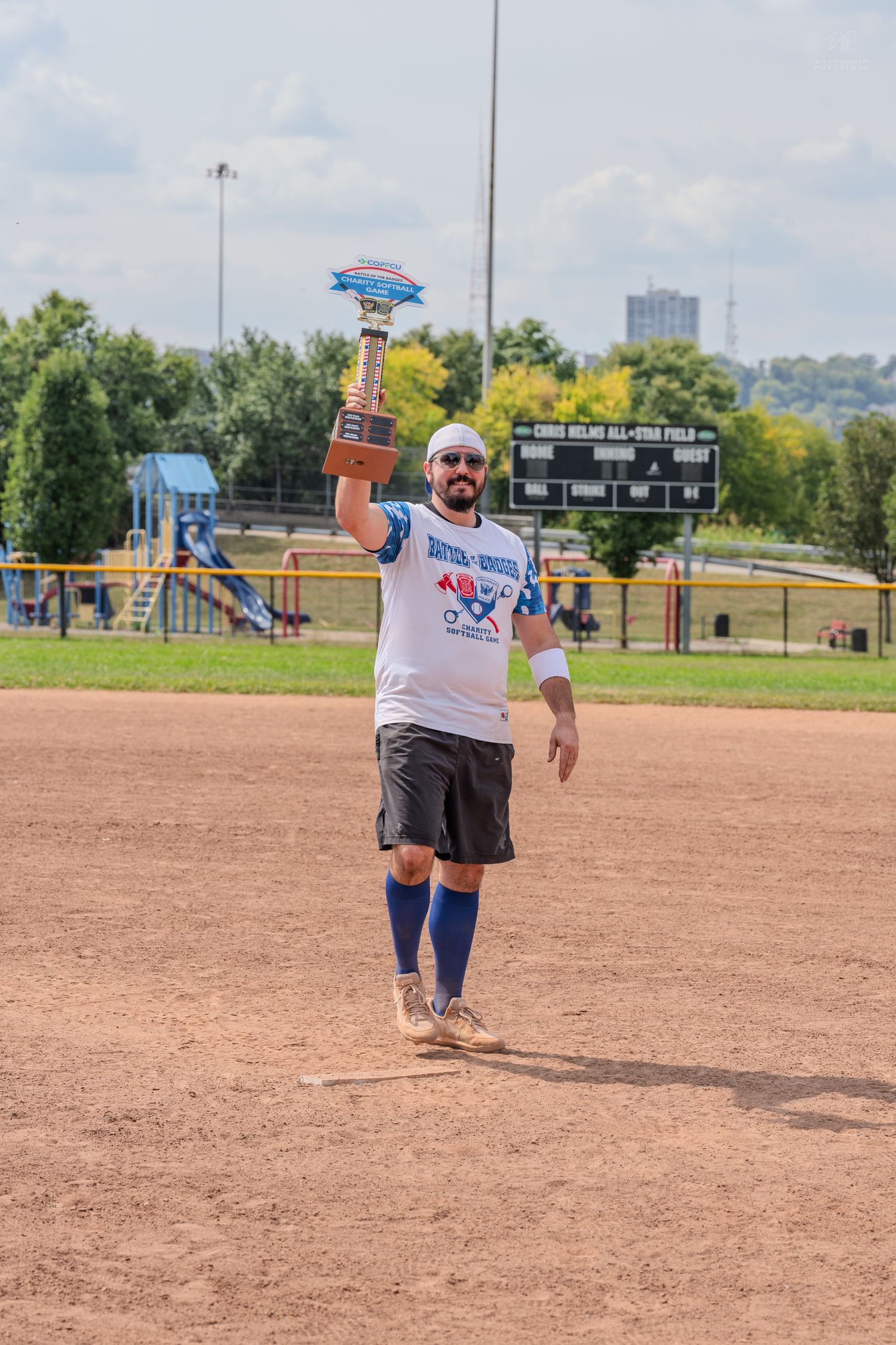 A bearded man in a white softball jersey raises a championship trophy overhead on a dirt baseball field.