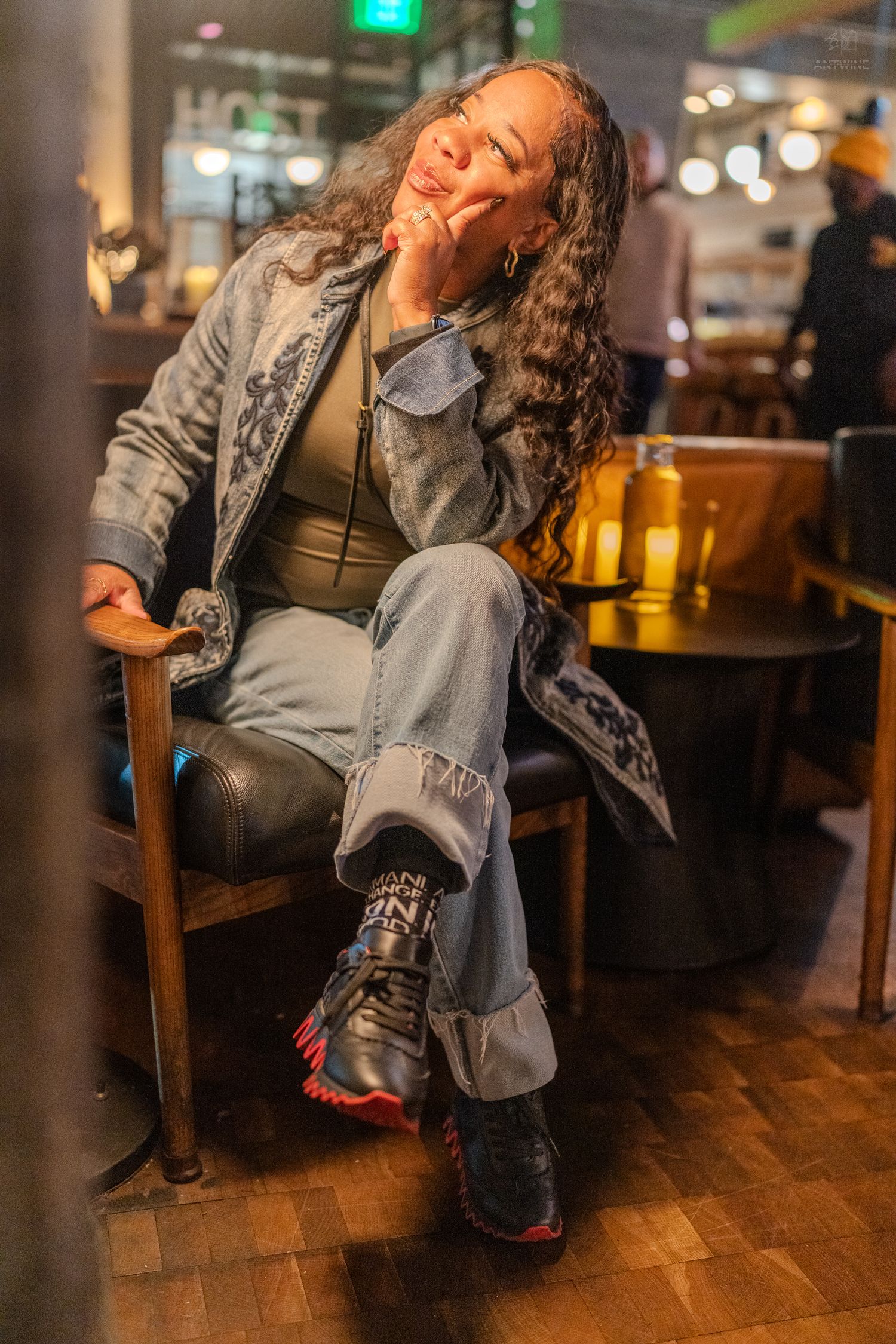 A stylish woman in denim poses thoughtfully in a leather chair at a warmly lit restaurant.