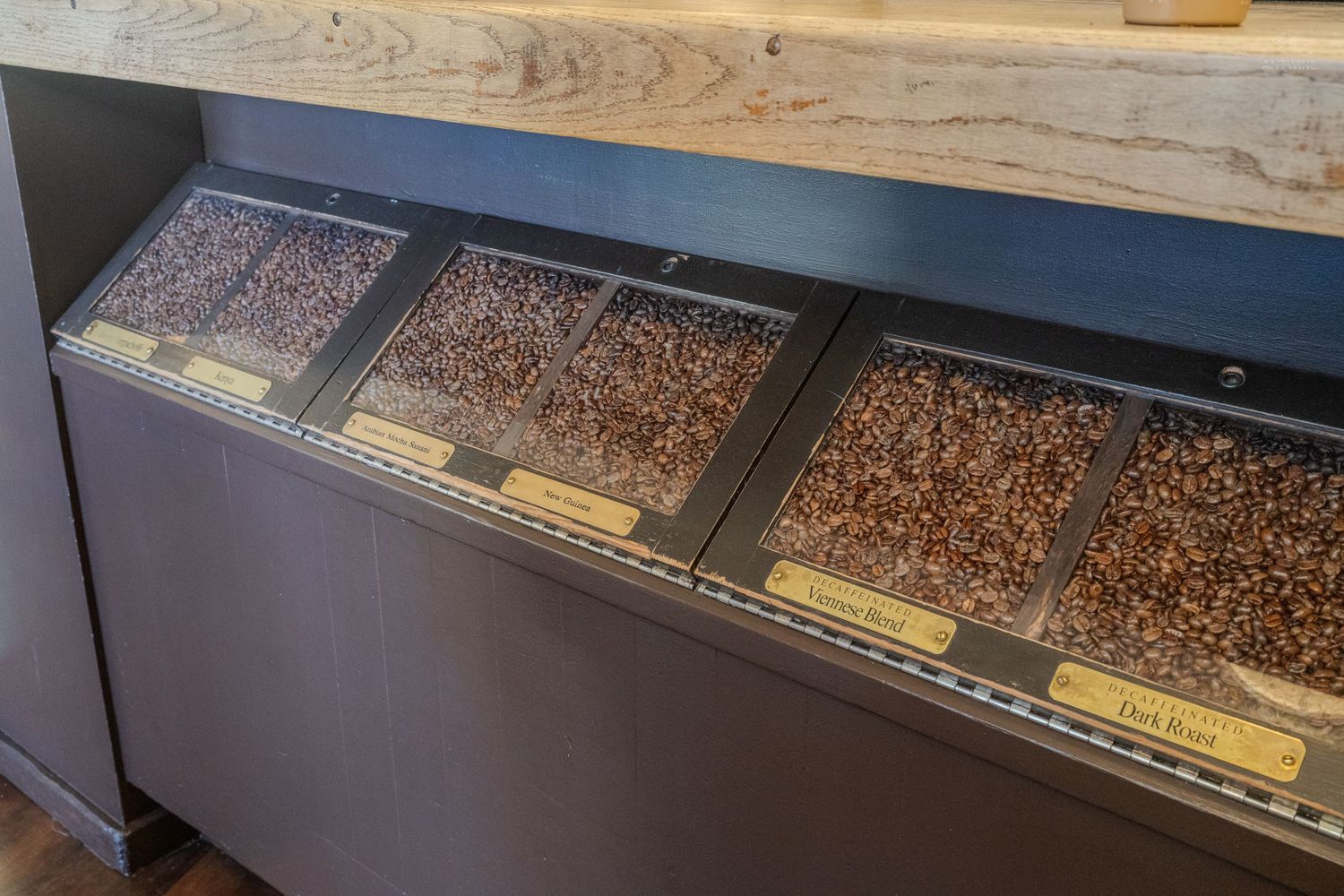 Three bins of assorted coffee beans on display at a coffee shop counter with yellow label tags.