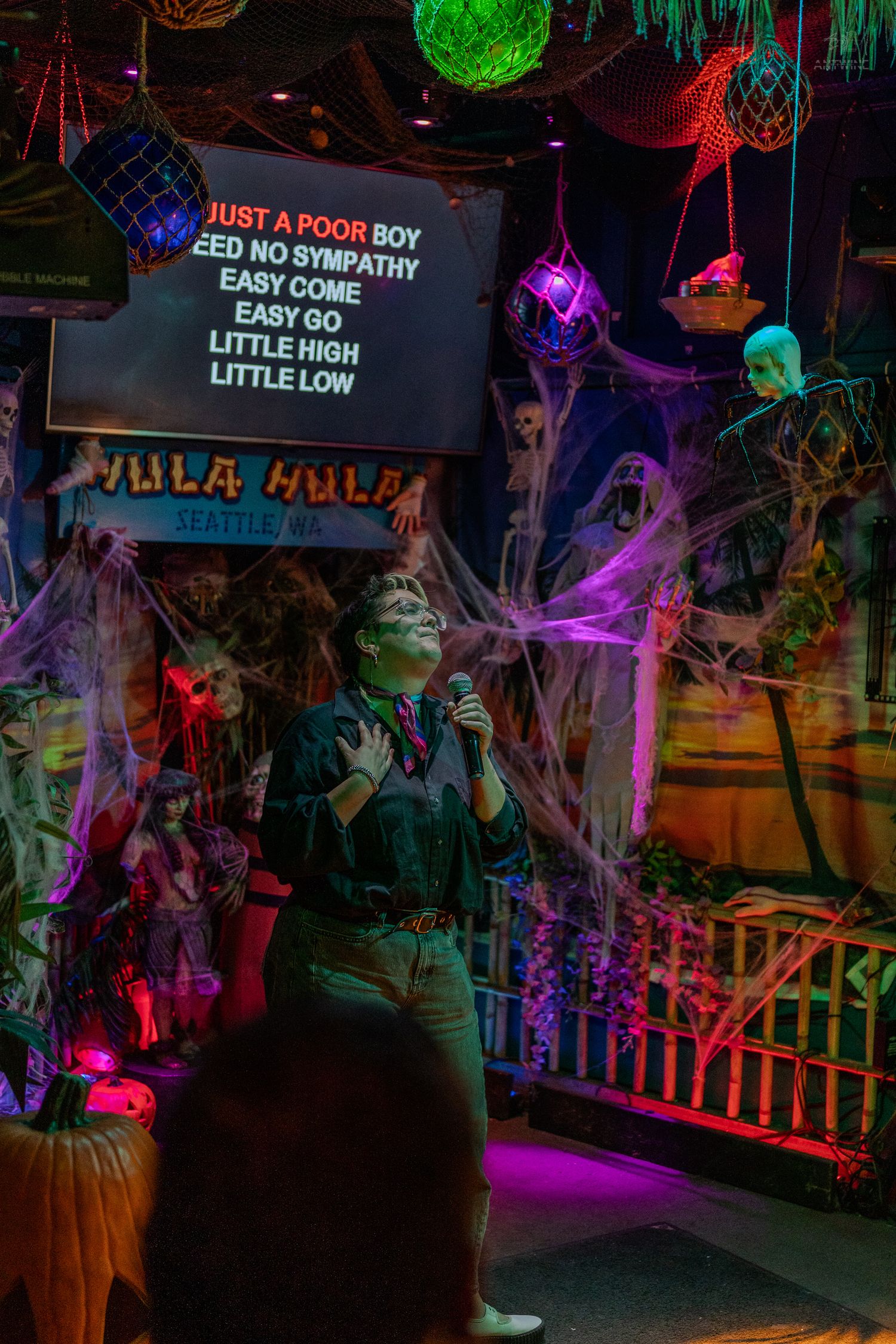 Person singing karaoke at Hula Hula Seattle bar surrounded by Halloween decorations and colorful lights.