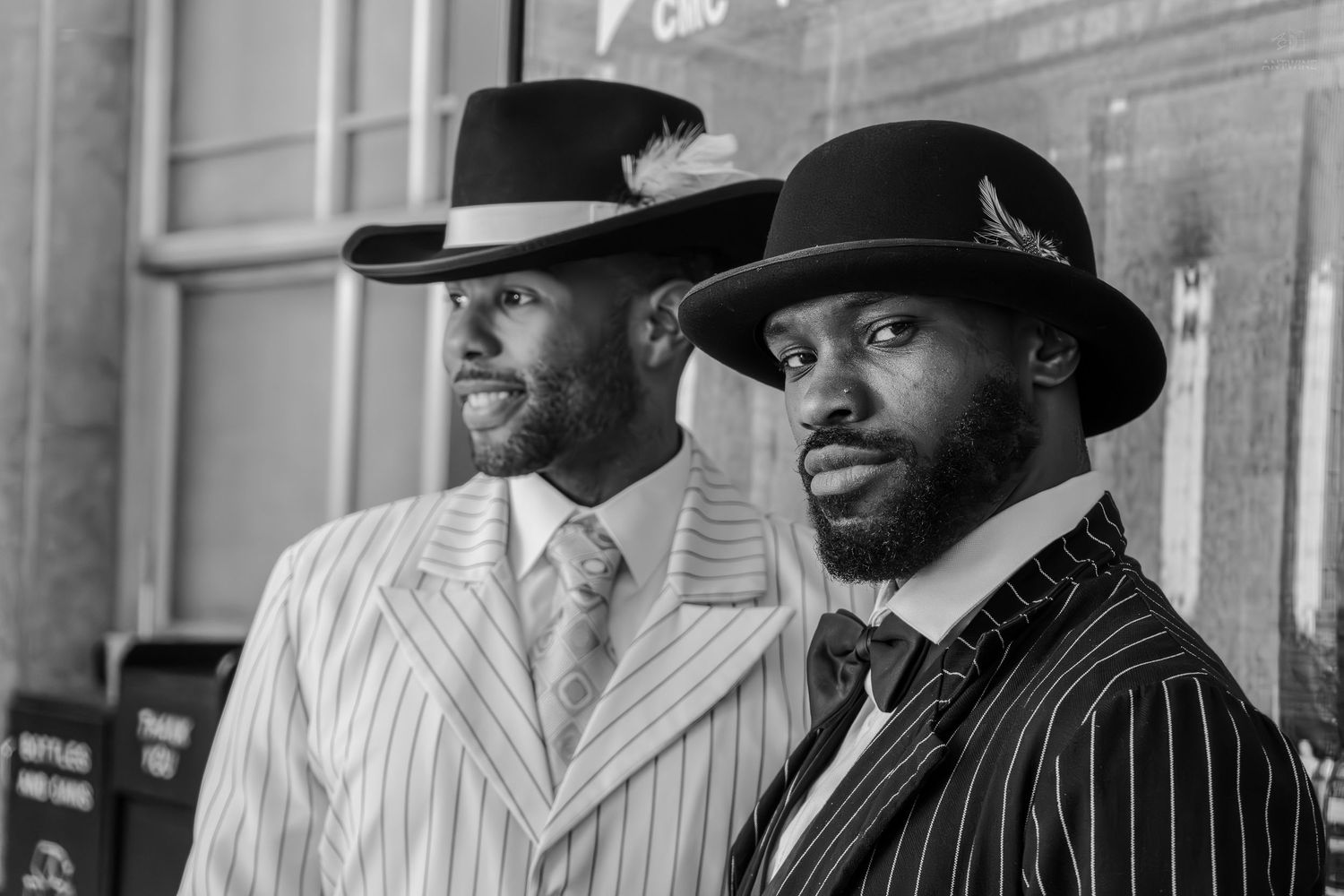 Two stylish men in pinstripe suits and wide-brim hats pose confidently in a classic black and white portrait photo.