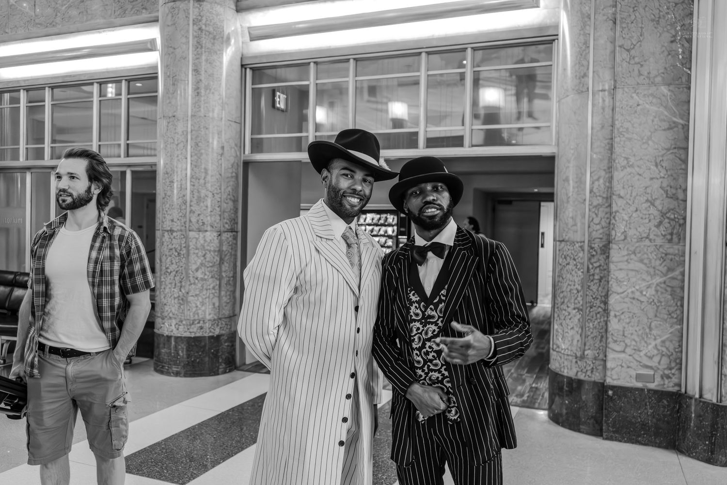 Two stylishly dressed men in pinstripe suits and hats pose indoors in a black and white photo.